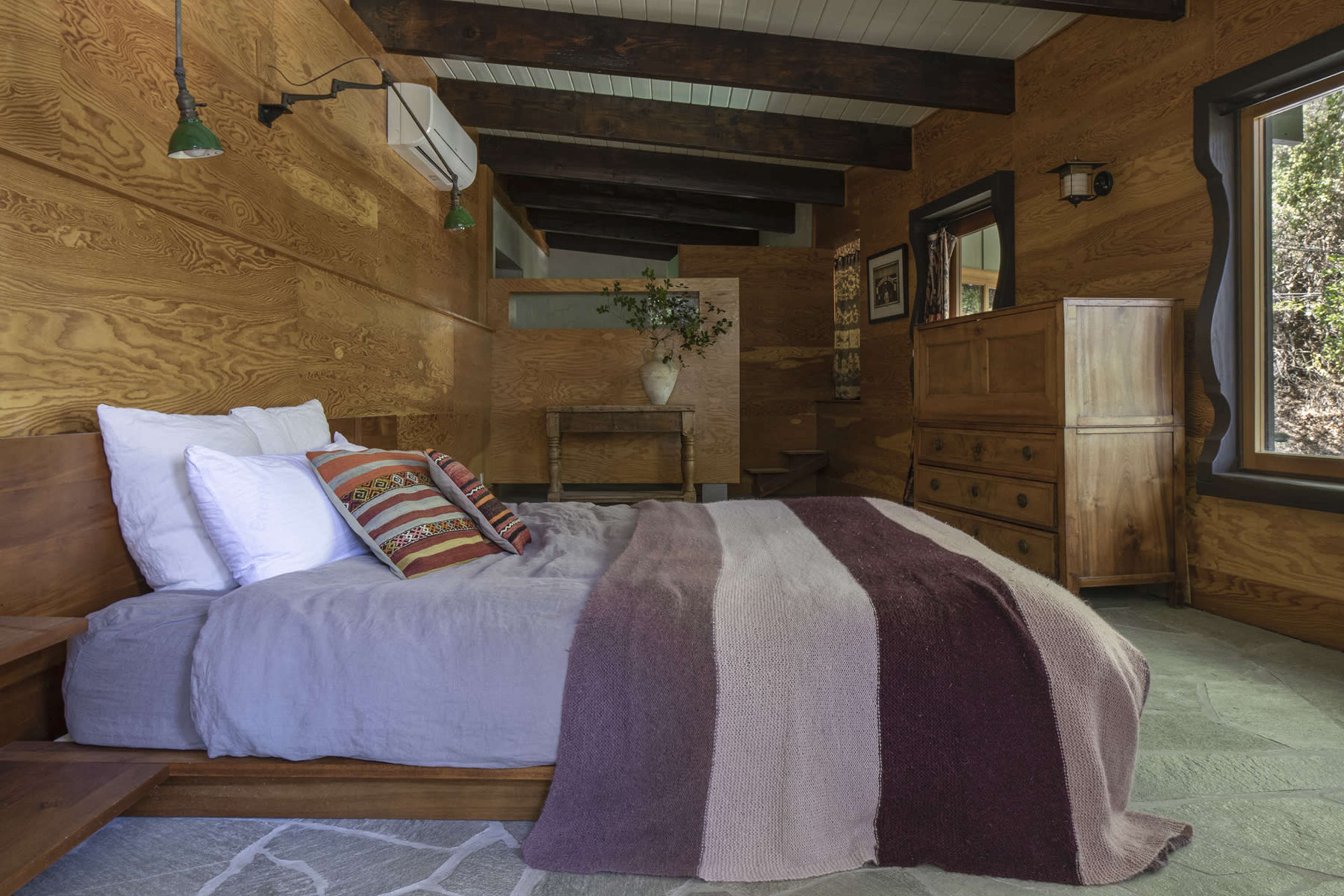 A rustic bedroom with wooden walls, a bed covered in a striped blanket, and a wooden dresser and table in the background.