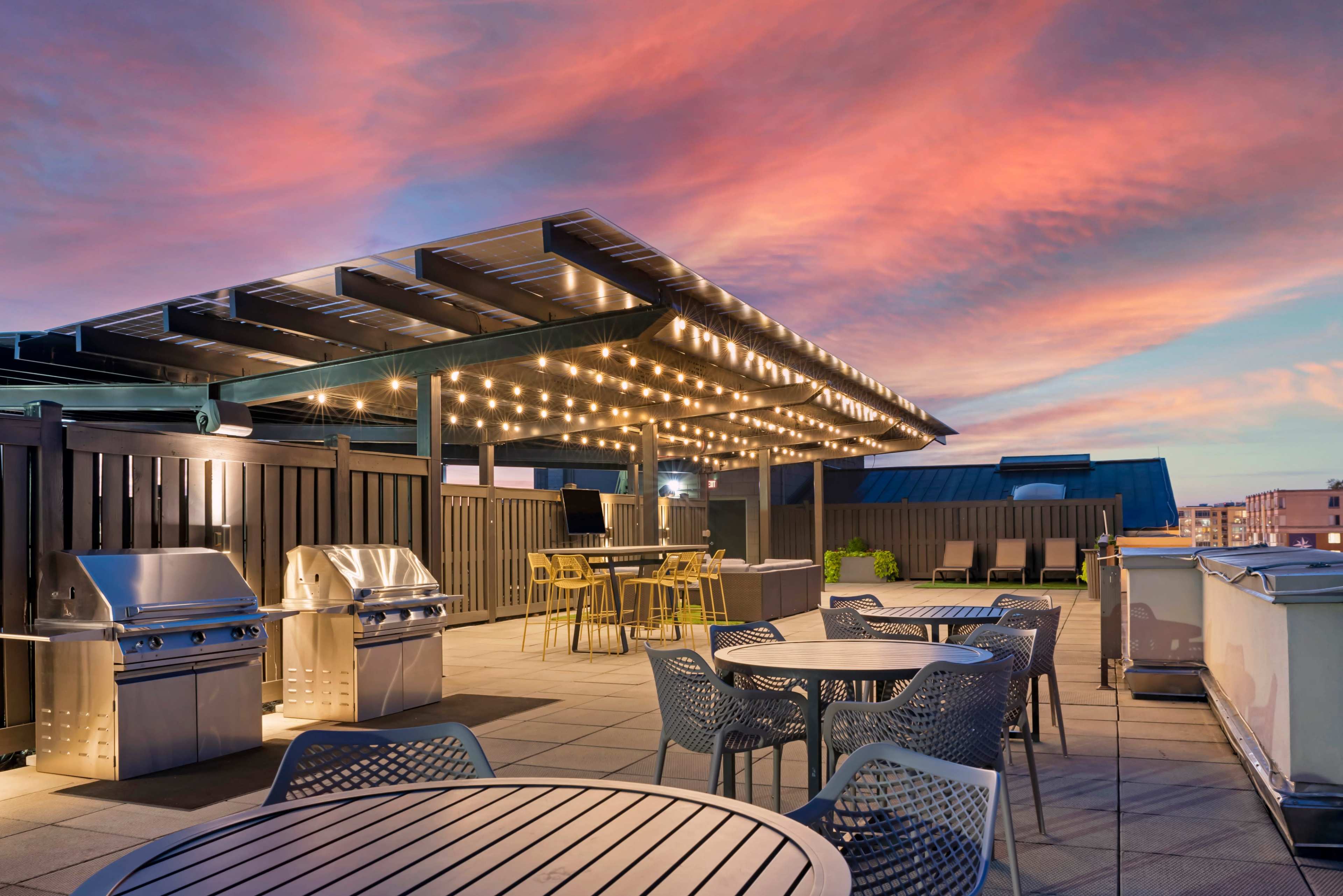 A rooftop patio features outdoor grills, dining tables, and string lights under a vibrant sunset sky.