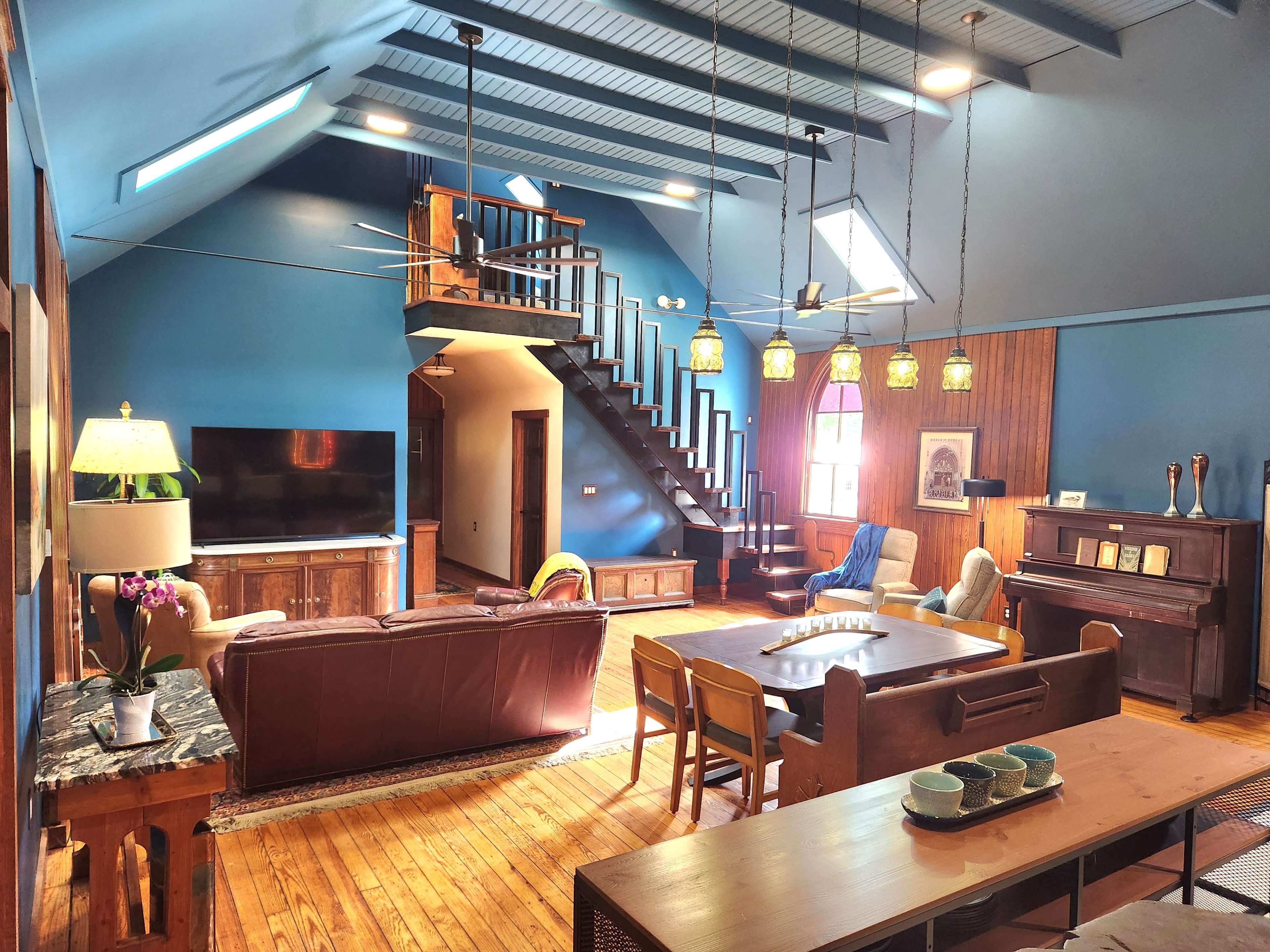 The image shows a spacious living area with blue walls, wooden flooring, a staircase, and various furniture pieces including a sofa, dining table, and a piano.