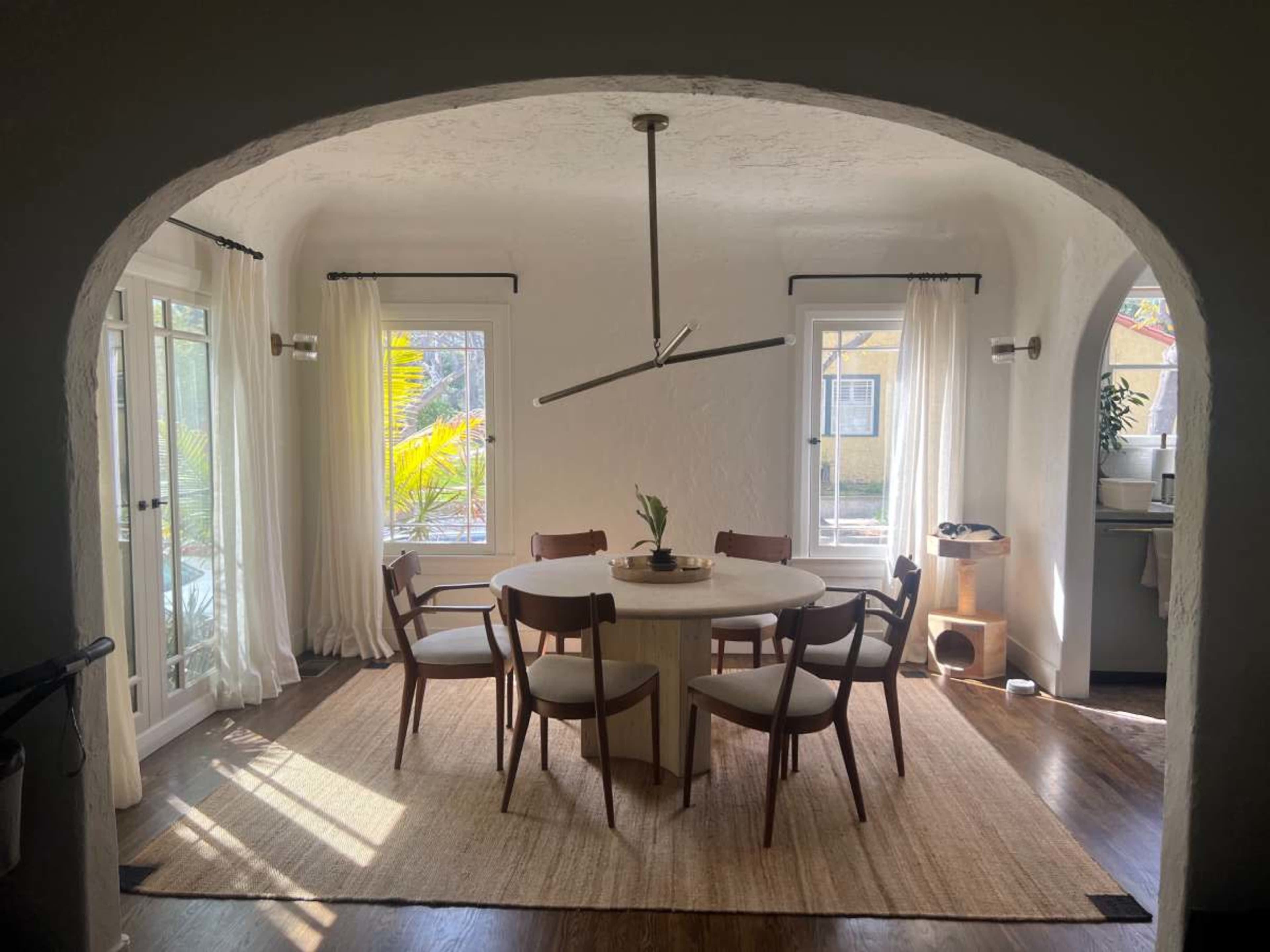 A round dining table surrounded by six wooden chairs is situated in a sunlit room with white walls and large windows, framed by light curtains.