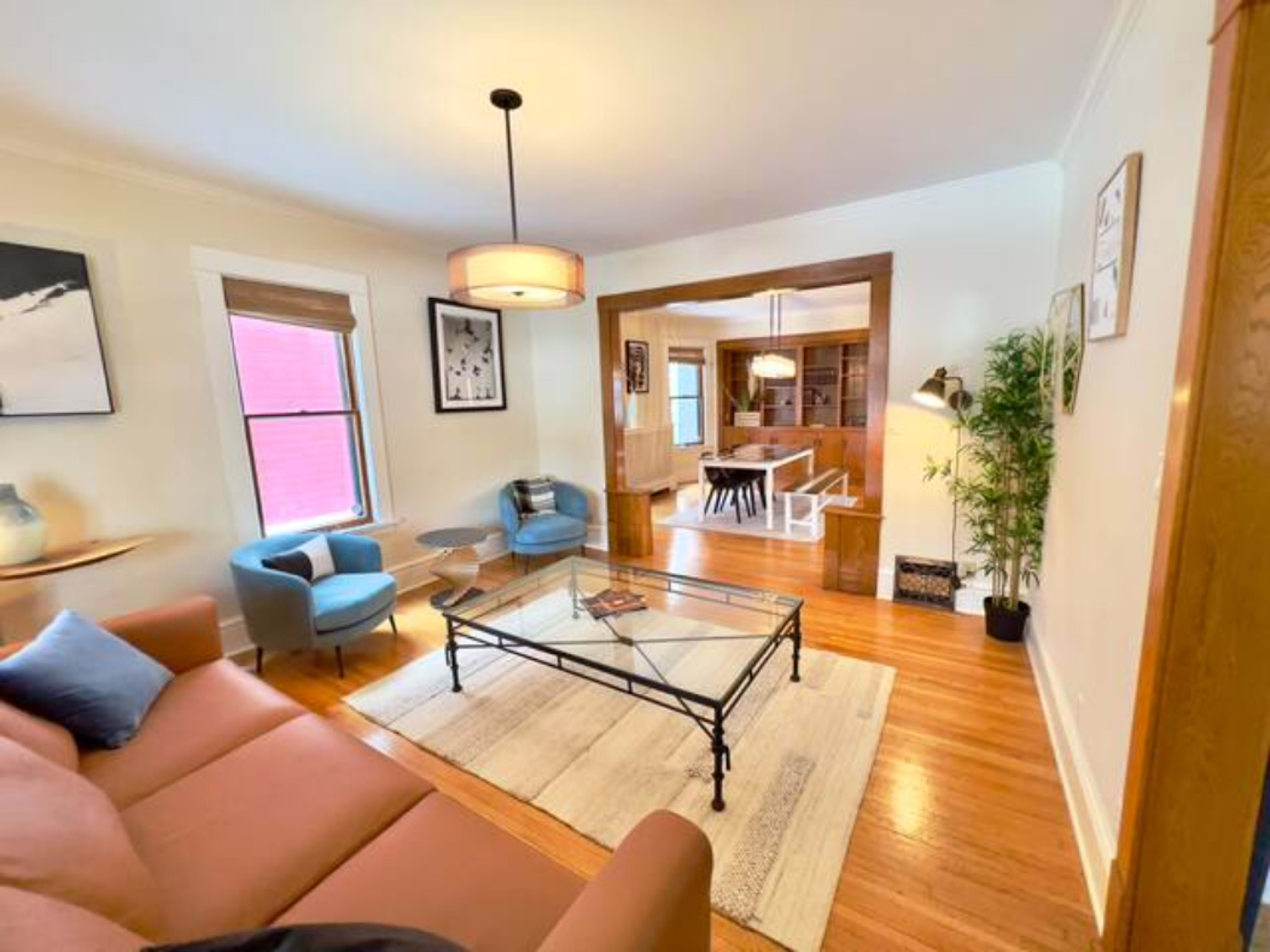 The living room features a brown sofa, a glass coffee table, and two blue accent chairs, leading to a dining area with wooden furniture.