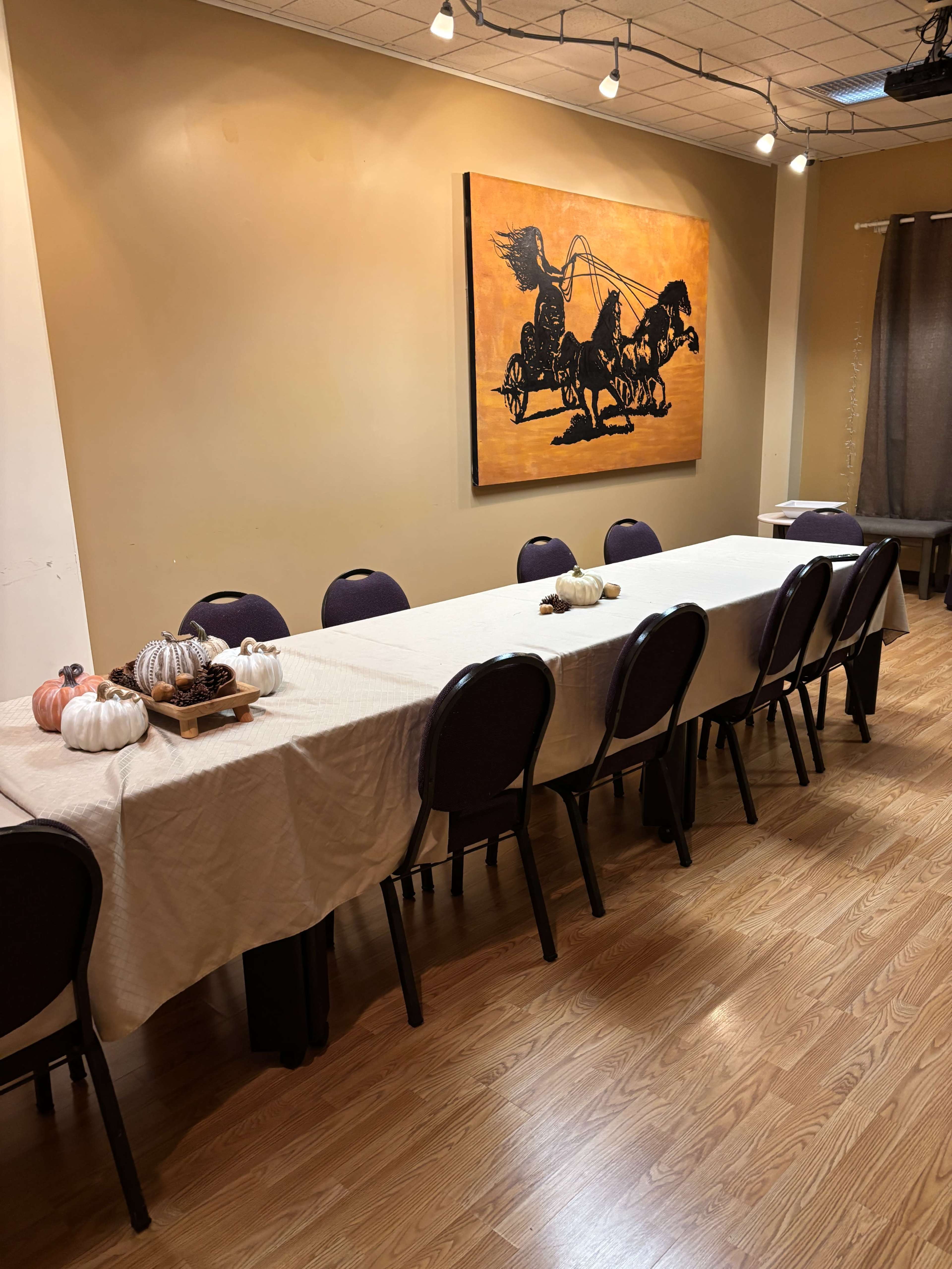 A long table covered with a light cloth, flanked by chairs, is set up in a room featuring a large painting of a horse-drawn carriage on the wall.