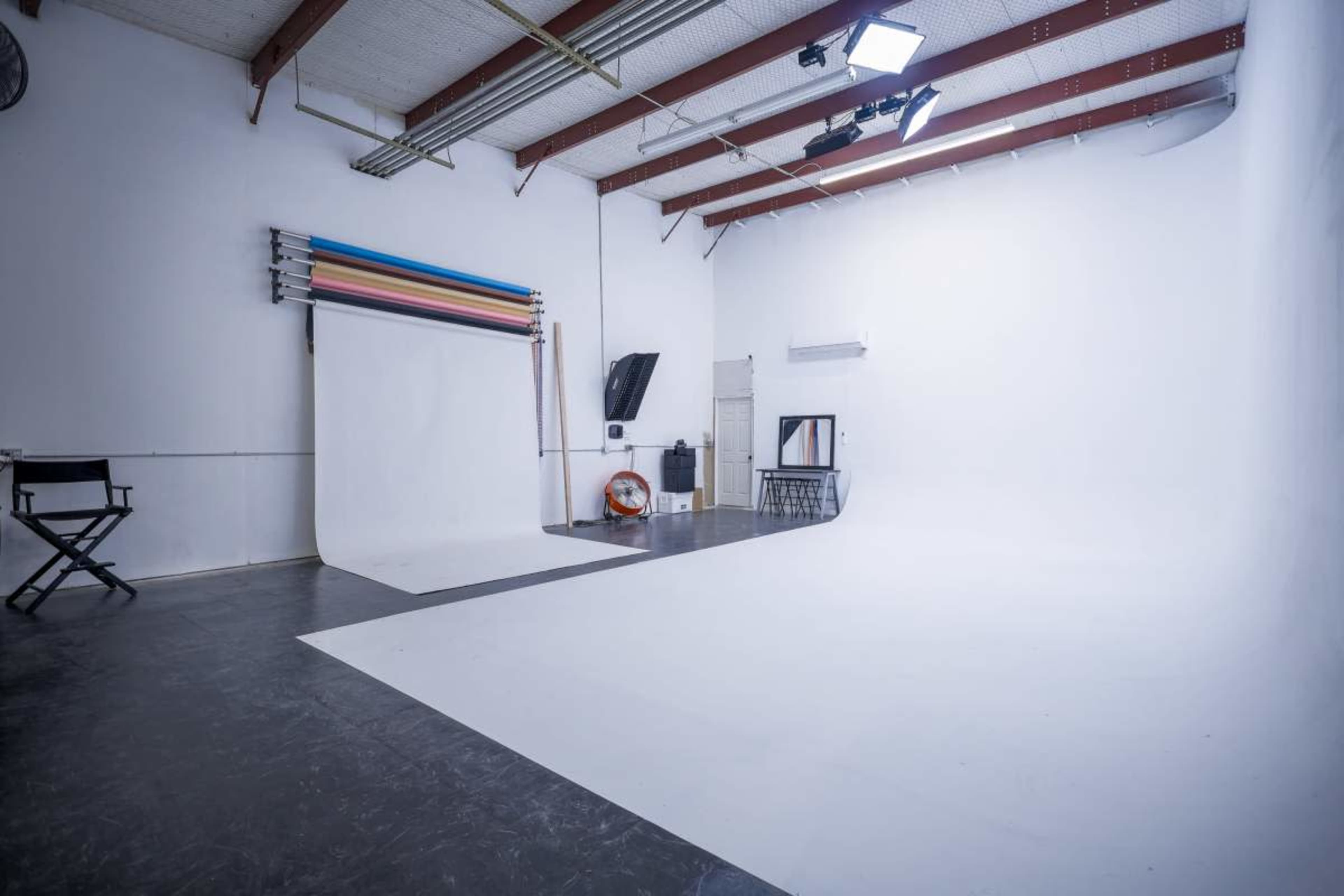 The image shows a spacious photography studio with white walls and a large white backdrop, along with a director's chair and various equipment in the background.