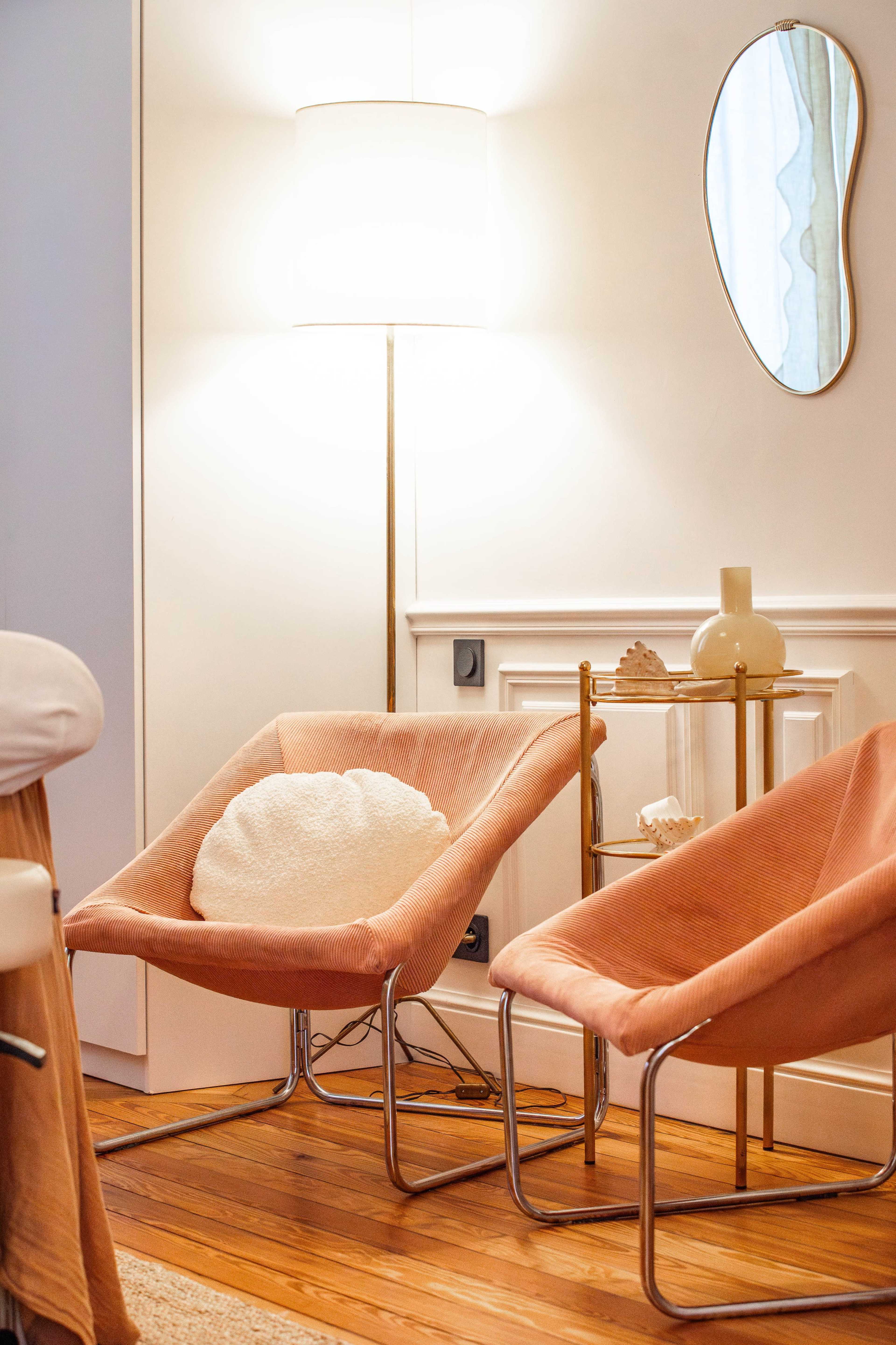 The image shows two modern chairs with a round pillow beside a lamp and a small table against a wall in a cozy interior space.
