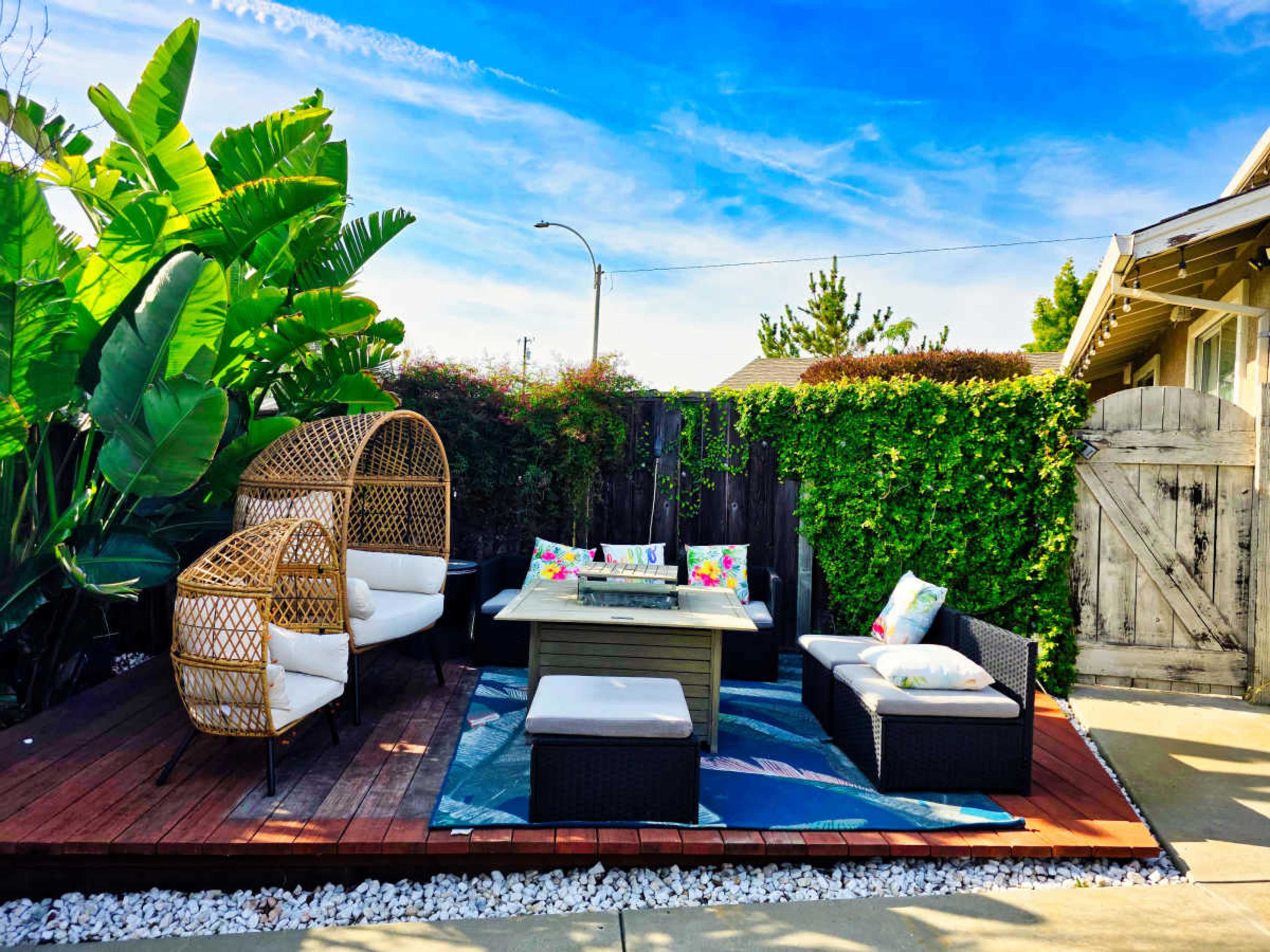 A lounge area with a wooden deck features two wicker chairs, a low table, and cushions surrounded by greenery and a wooden gate.