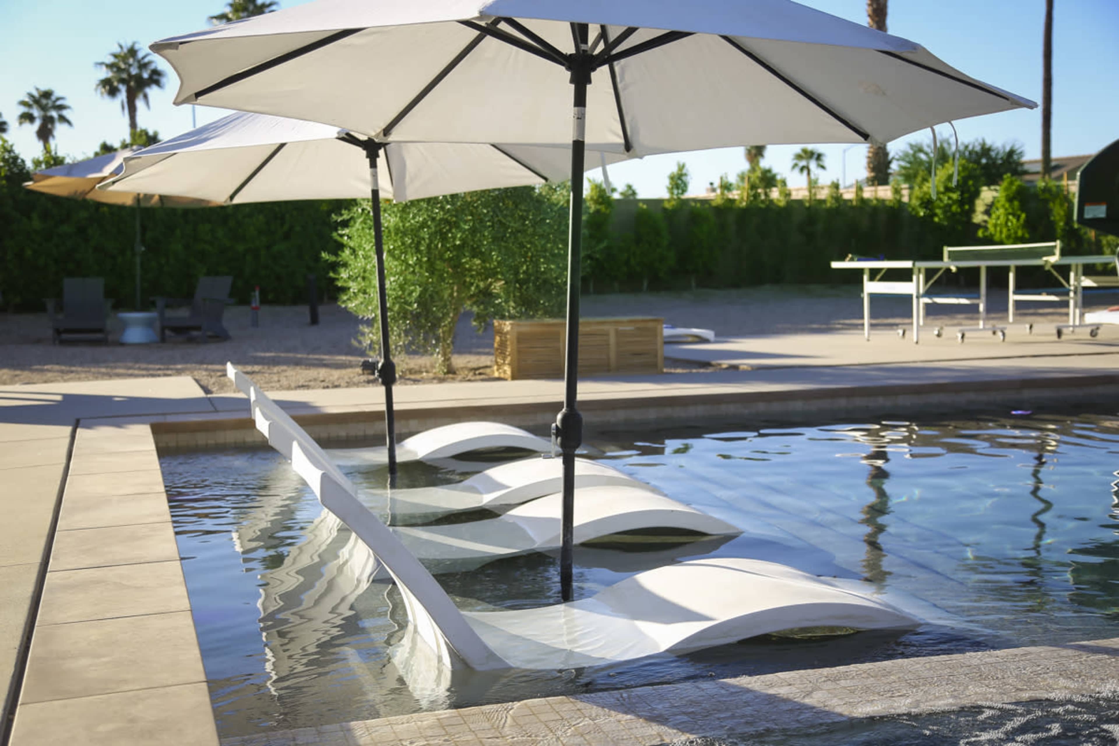 A swimming pool features white loungers partially submerged in the water, shaded by large umbrellas.