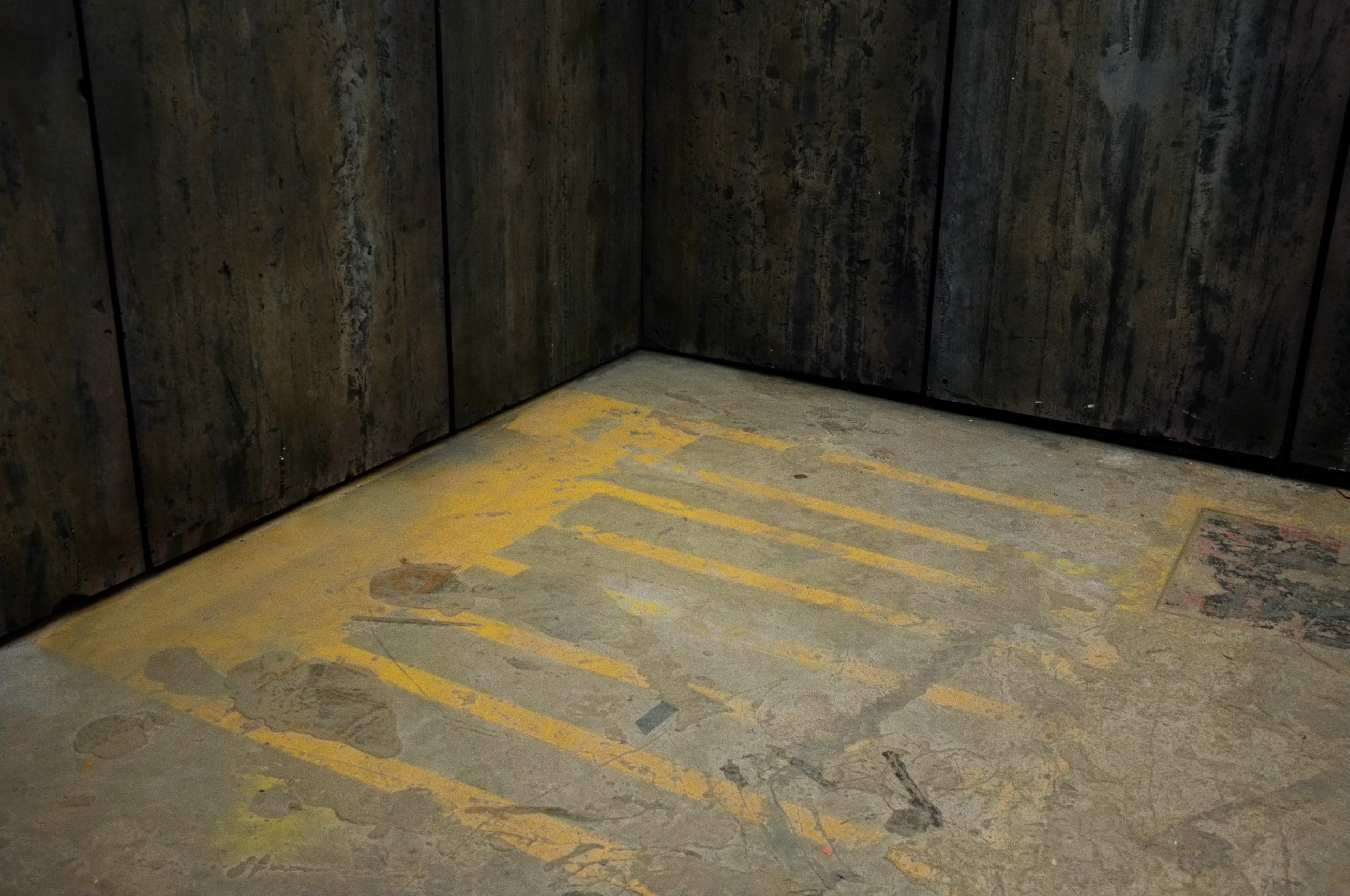 The image shows a corner of a dimly lit room with a concrete floor marked by yellow stripes and a worn rug.