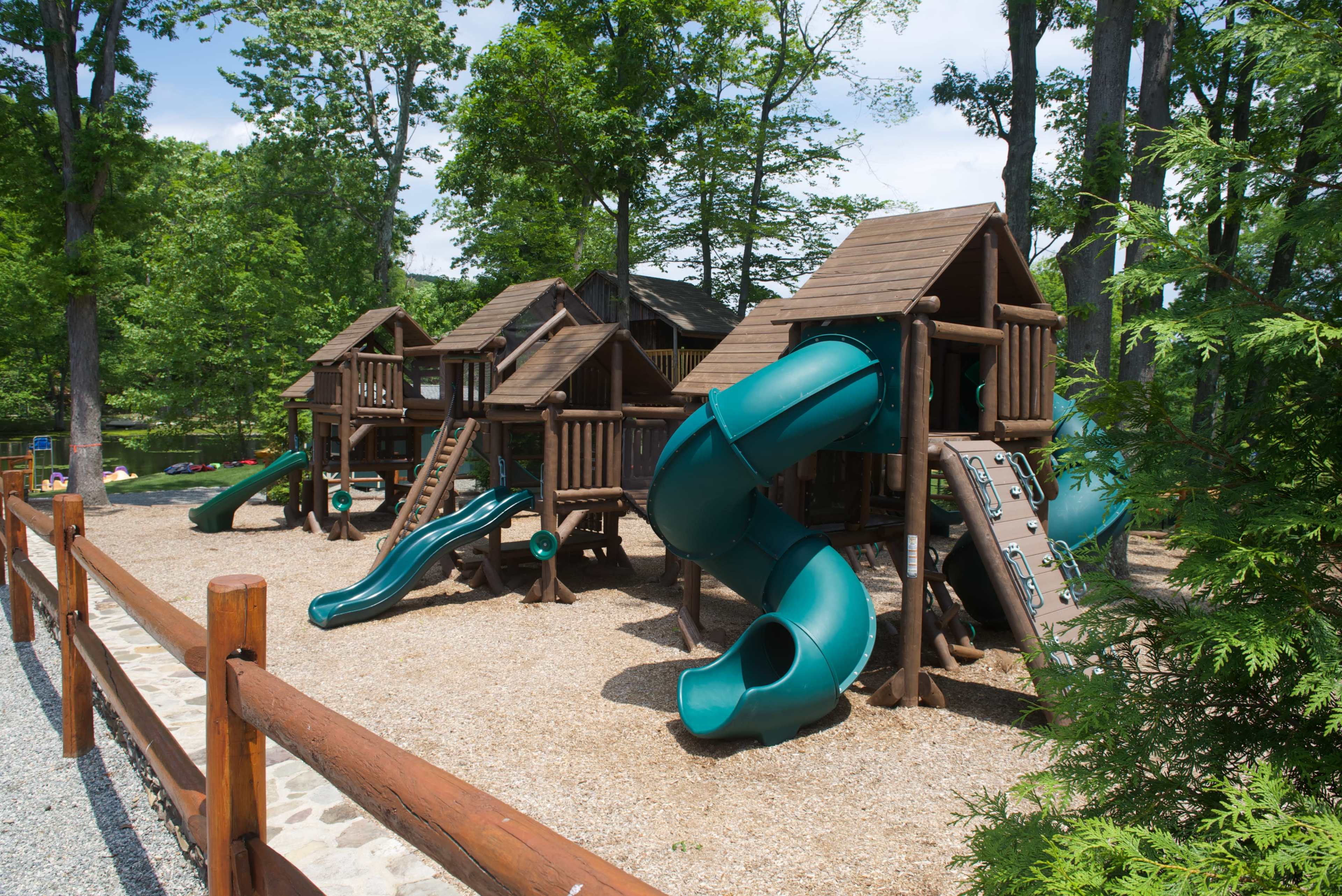 The image shows a wooden playground structure with two slides located in a shaded area surrounded by trees.