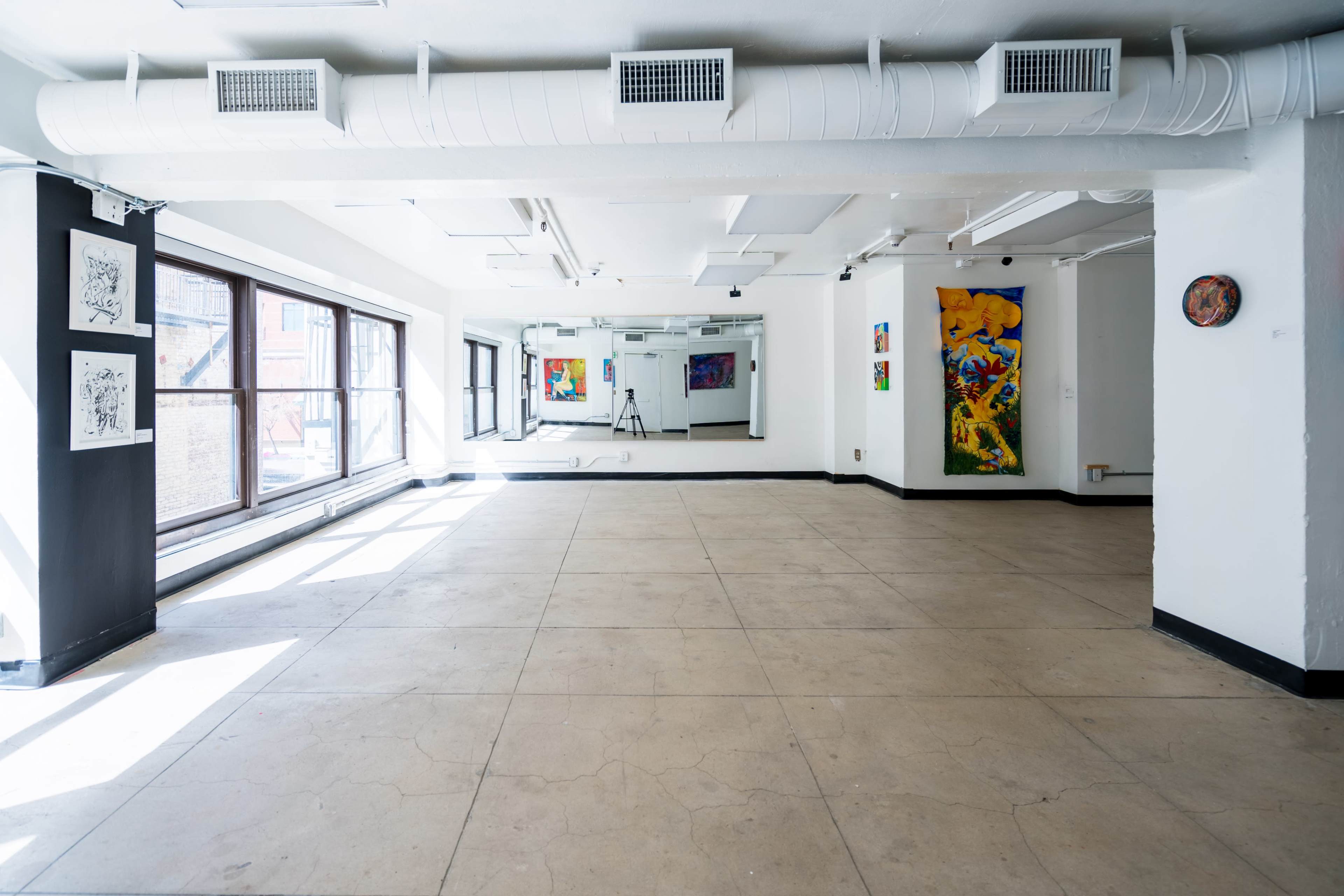 A spacious art gallery with large windows, showcasing colorful paintings on the walls and bright, empty flooring.