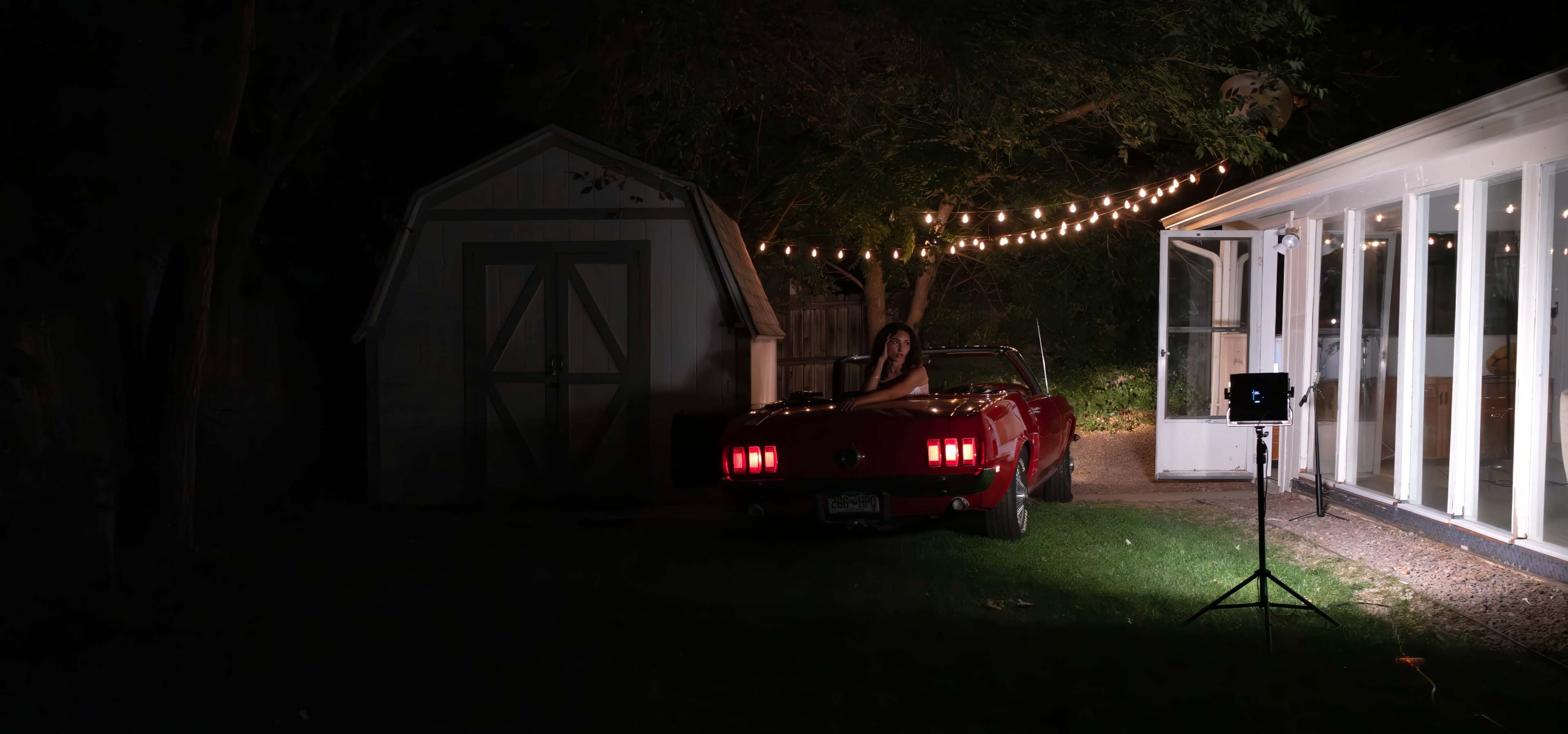 A red convertible car is parked next to a shed and a glass-walled structure, illuminated by string lights in a dark outdoor setting.