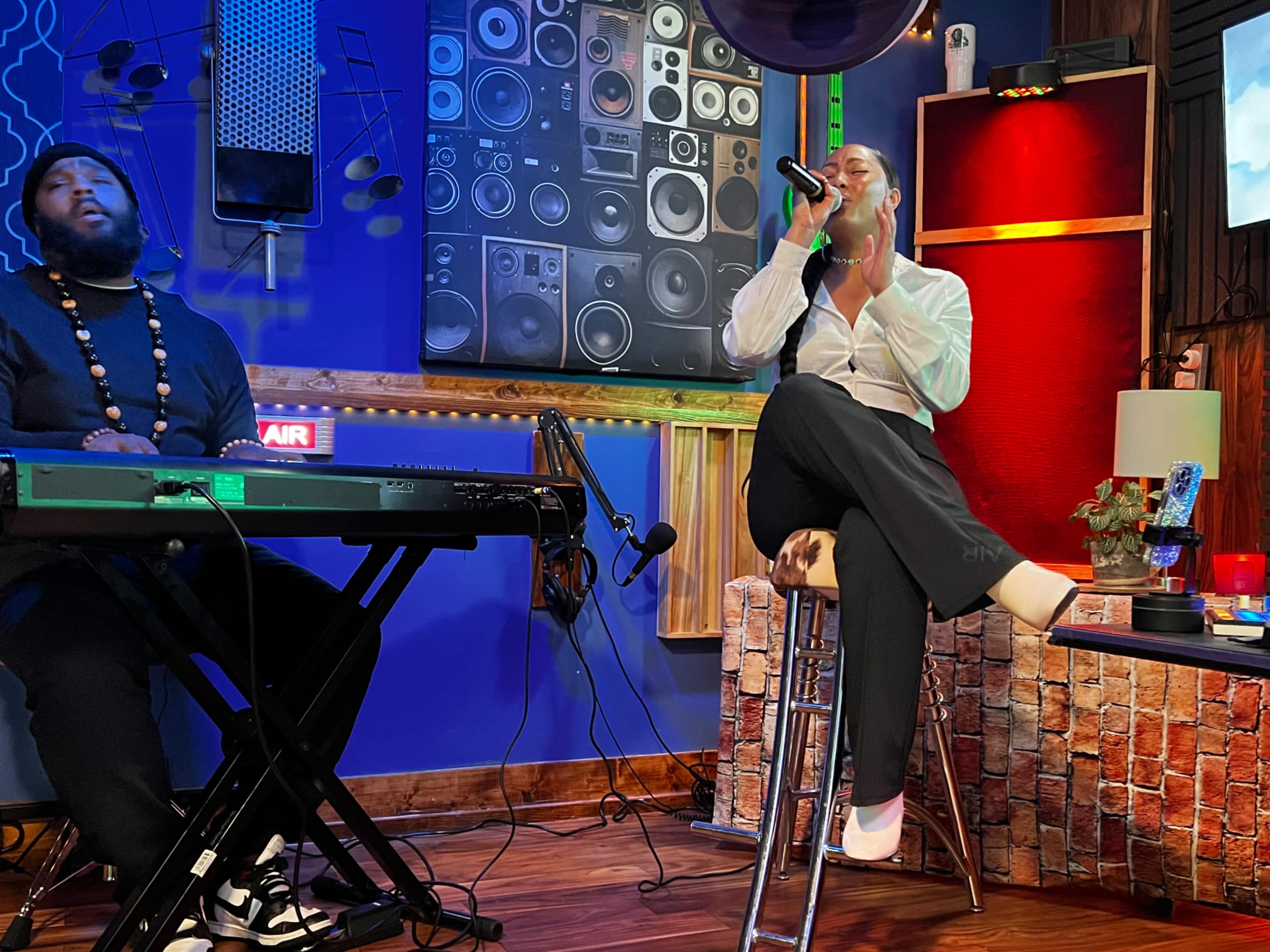 A performer sings into a microphone while sitting on a stool next to a keyboardist in a colorful music studio.