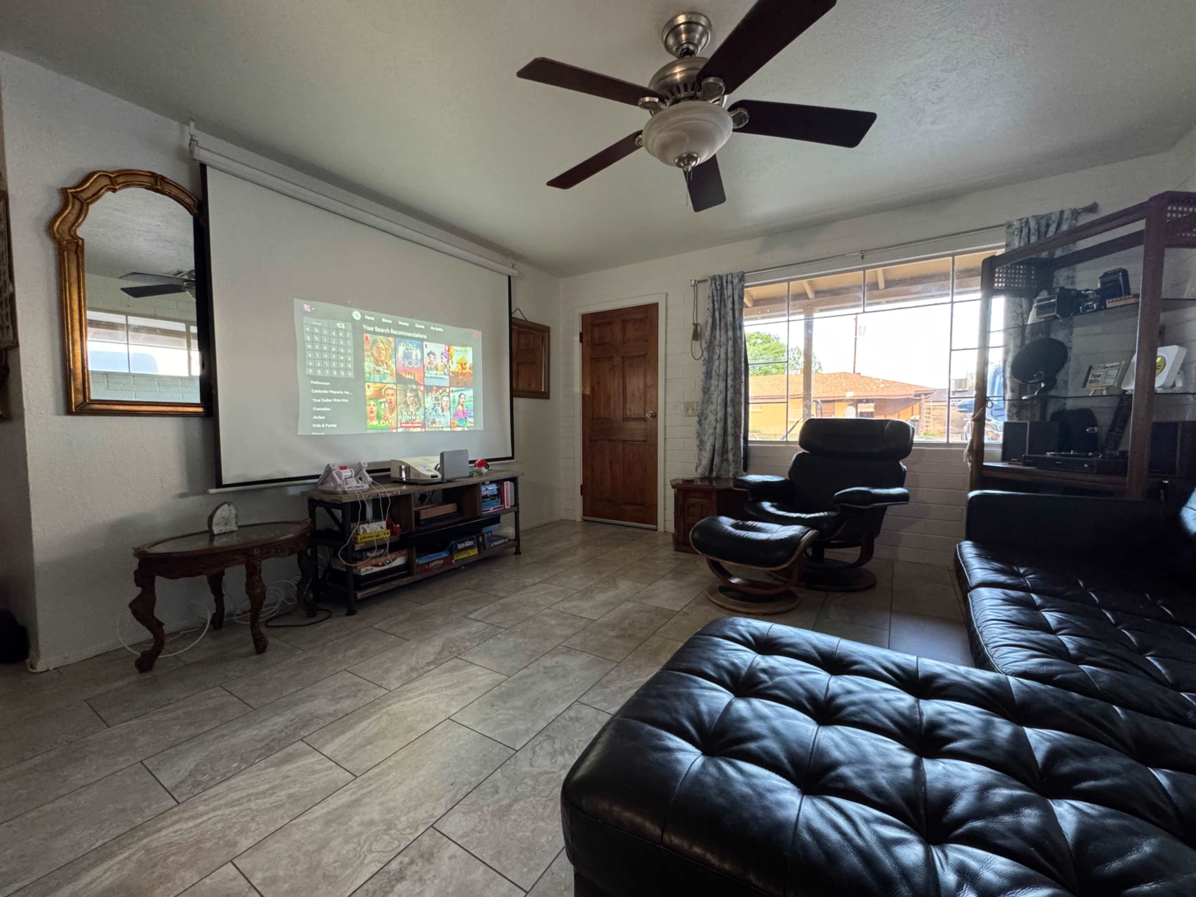 The image shows a living room with a projector screen displaying content, a black leather couch, and a wooden table with a fan overhead.