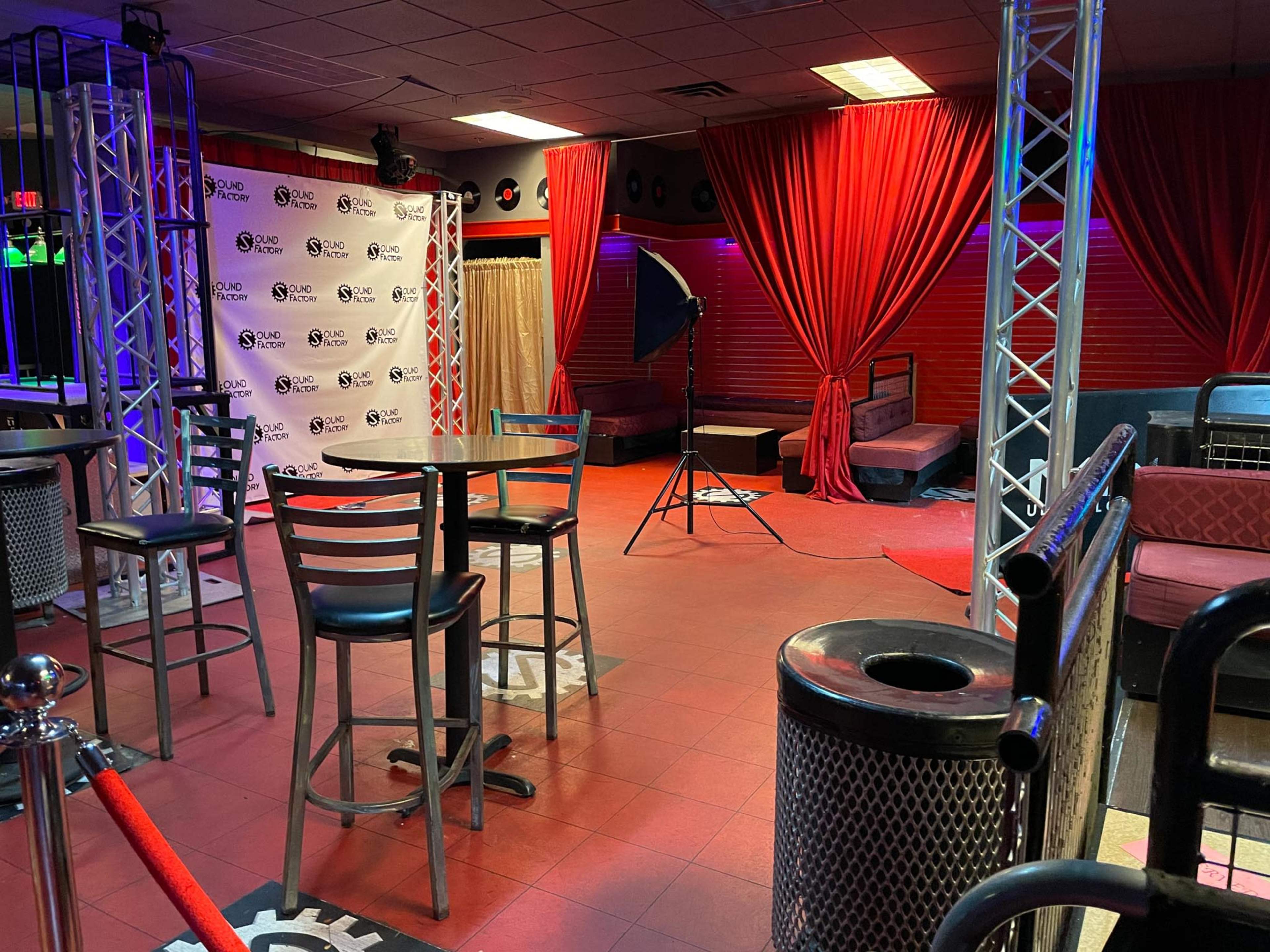 A dimly lit bar area with red curtains, empty tables, and a photography setup in the corner.
