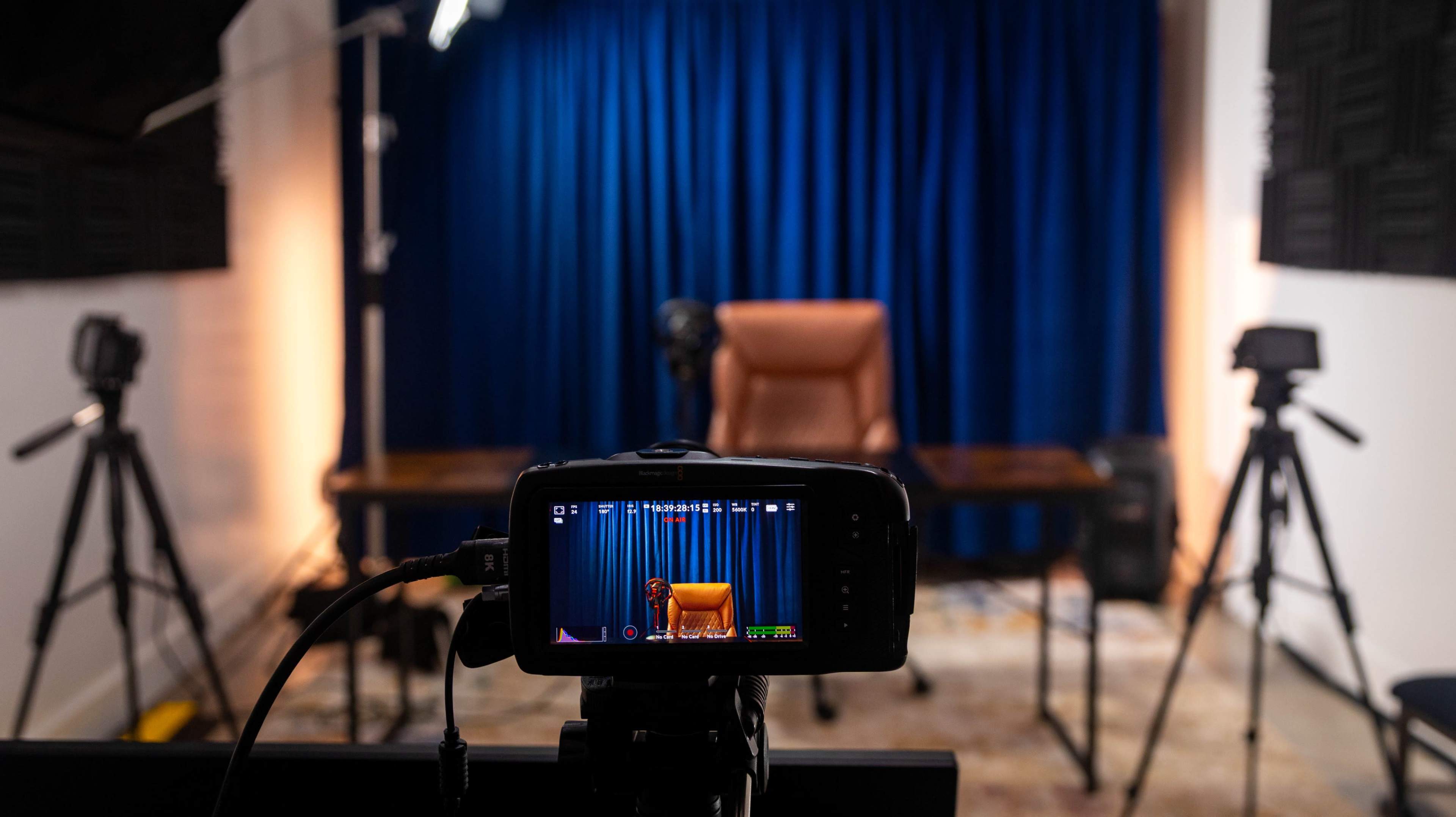 A camera is focused on a wooden desk and an empty chair in front of a blue curtain, with additional cameras on tripods positioned nearby.