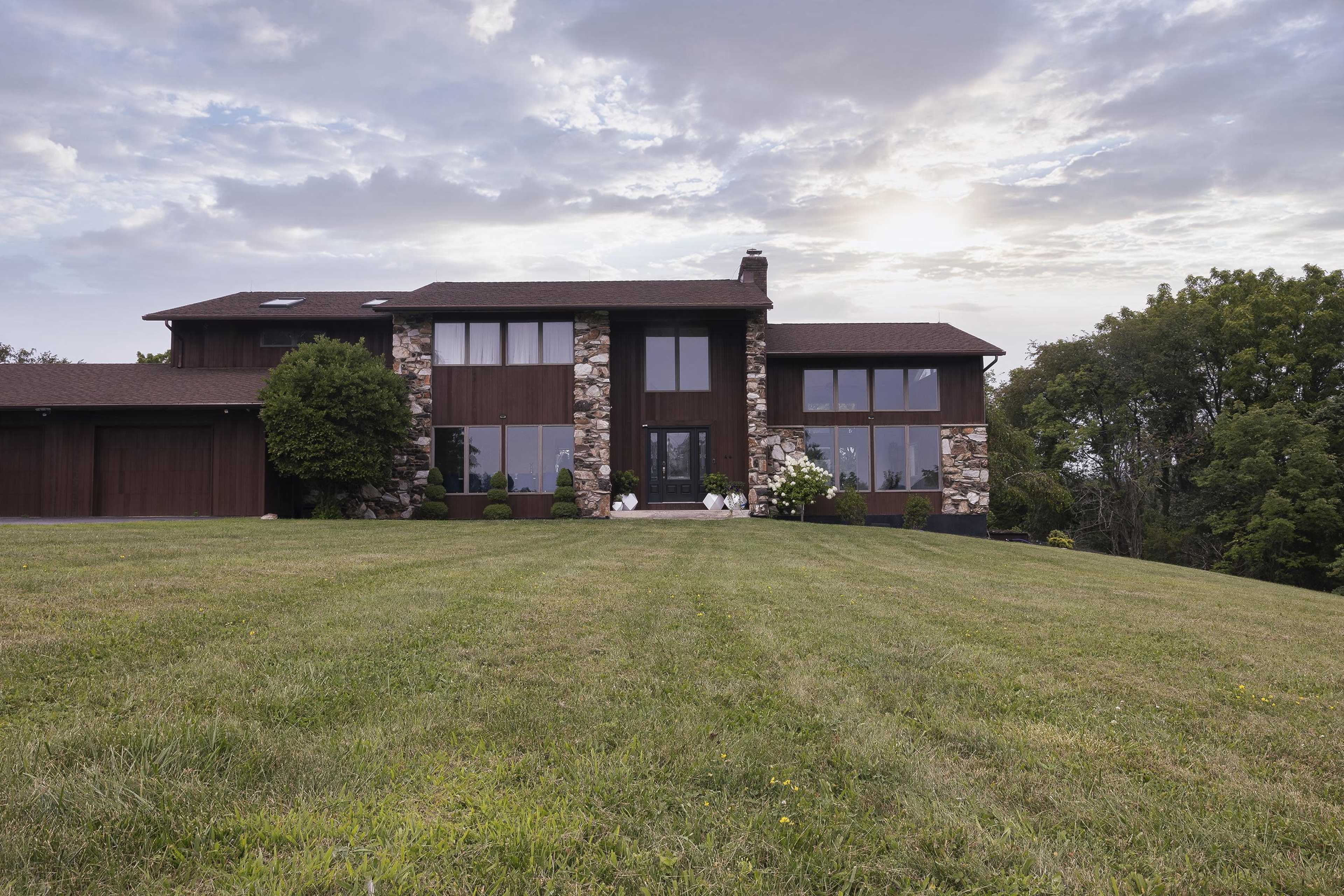 A modern house with a stone facade and large windows sits on a green lawn under a cloudy sky.