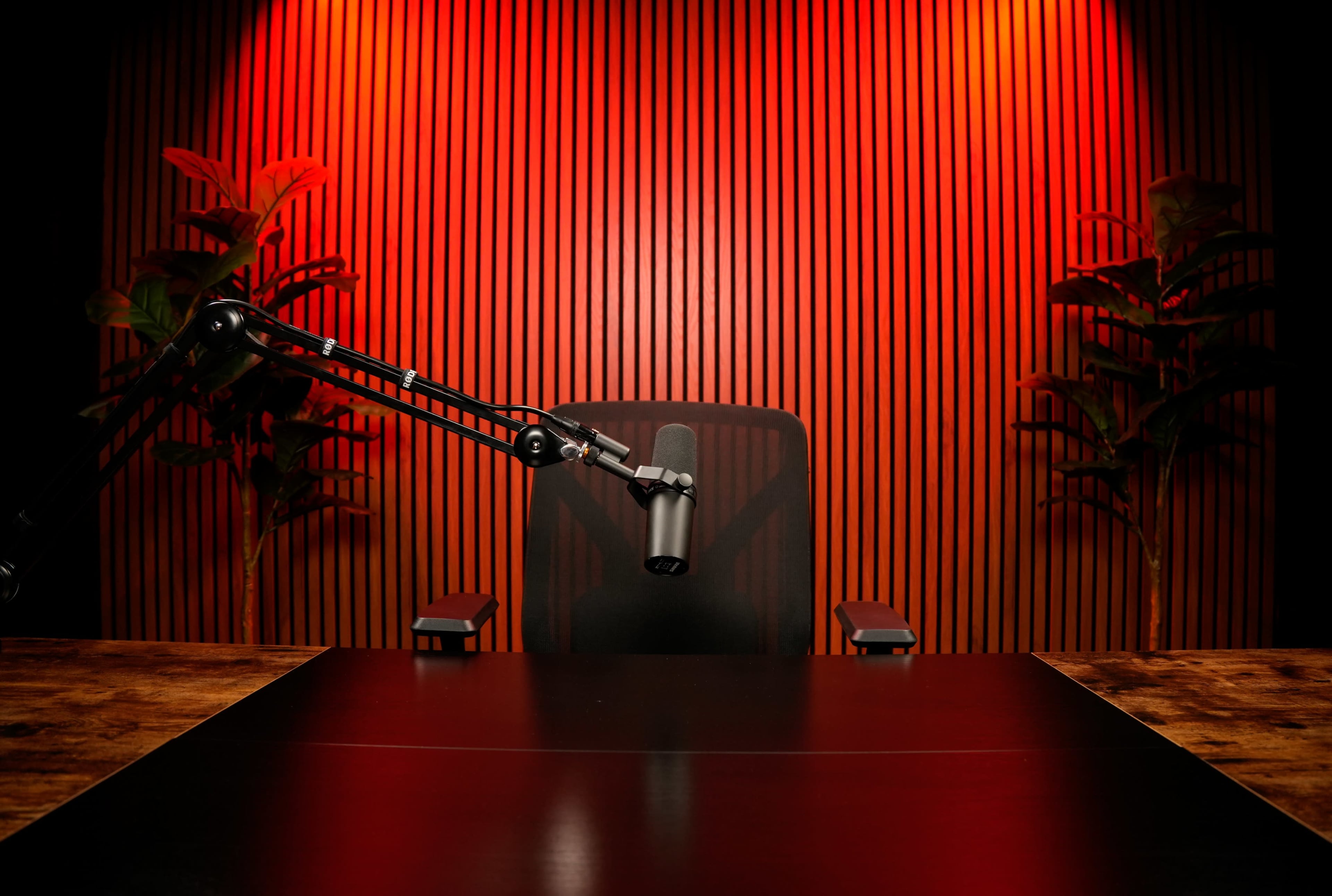 A microphone is positioned on an adjustable arm above a black table in front of an empty chair, with wooden paneling and red lighting in the background.