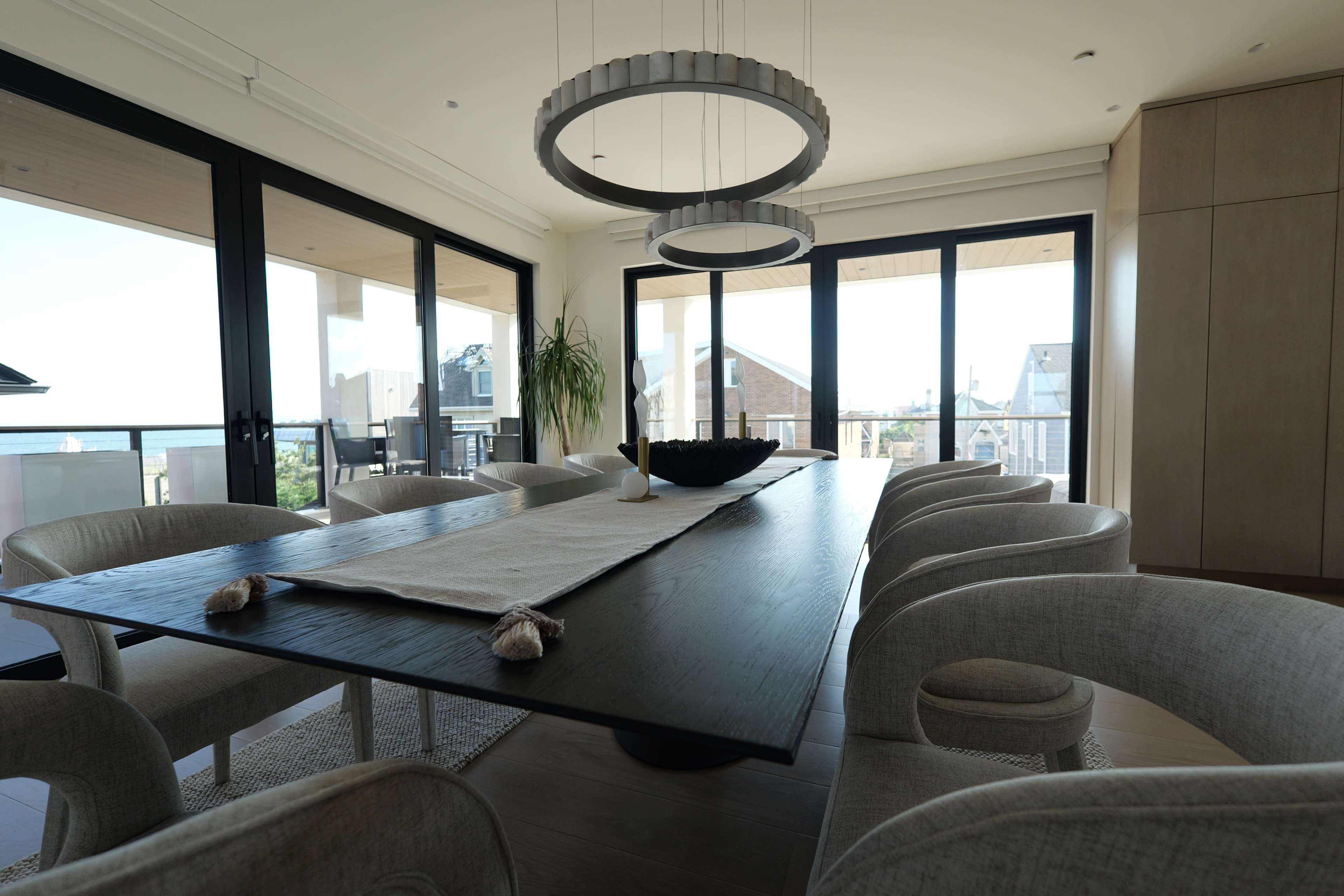 A modern dining room features a large black table surrounded by eight light-colored chairs, with large windows providing a view of the outdoors.