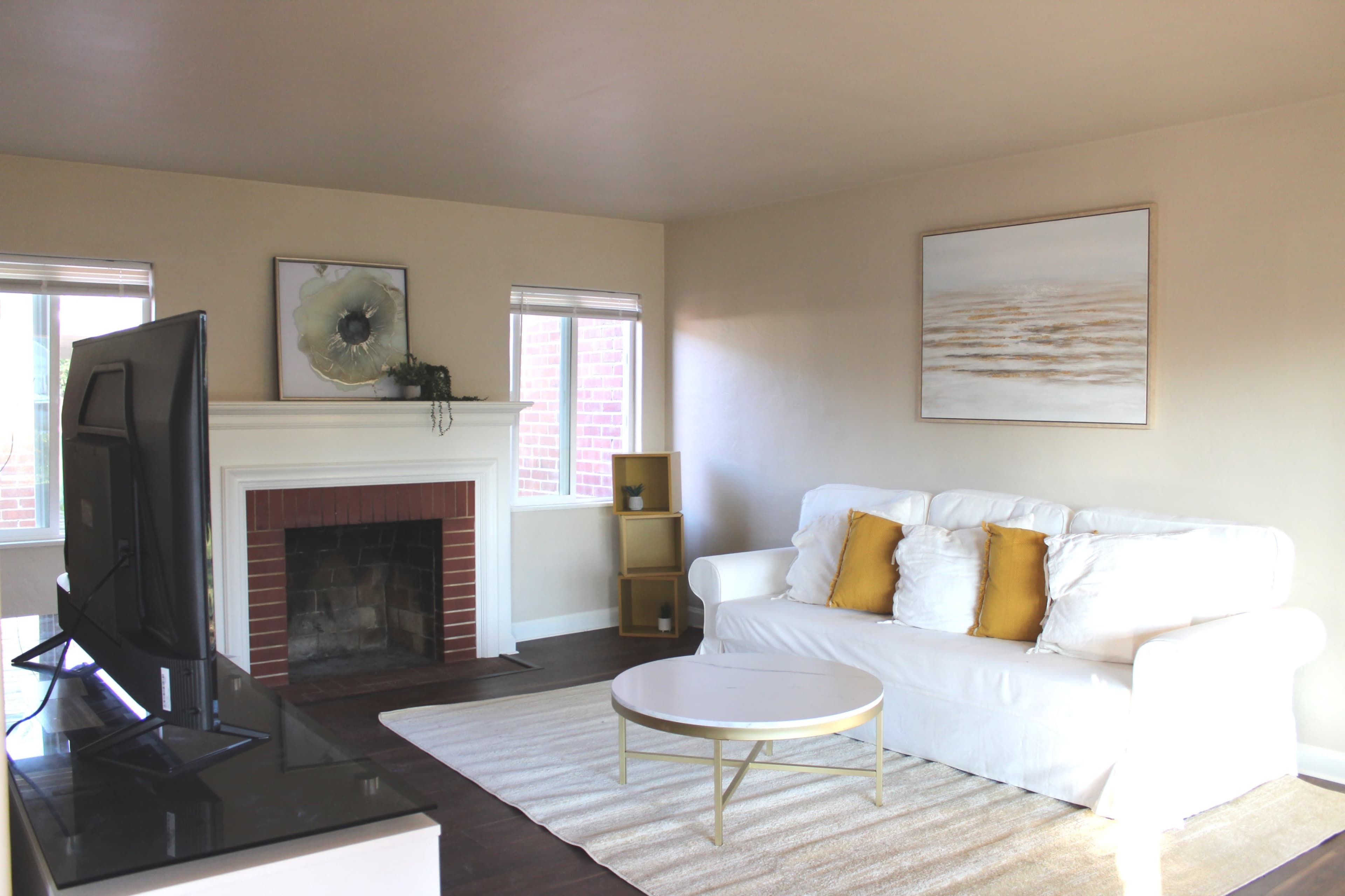 The image shows a living room with a white sofa, yellow pillows, a coffee table, a fireplace, and a large painting on the wall.