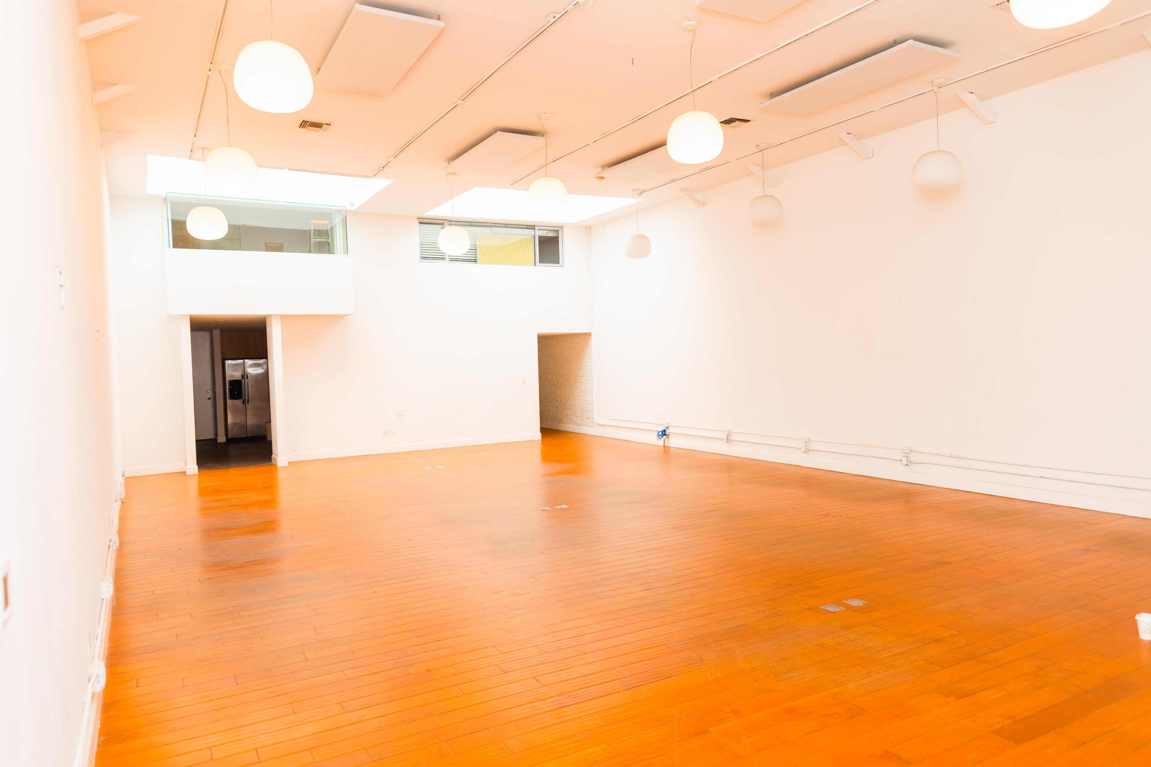 The image shows a spacious, empty room with wooden flooring, white walls, and several pendant lights hanging from the ceiling.