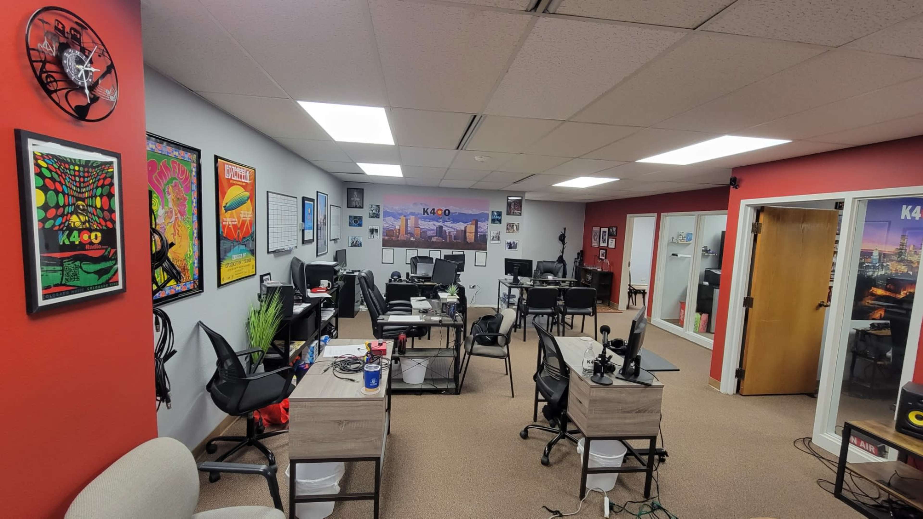 The image shows an office space with multiple desks, chairs, and decorative posters on the walls, along with a recording area visible through glass doors.