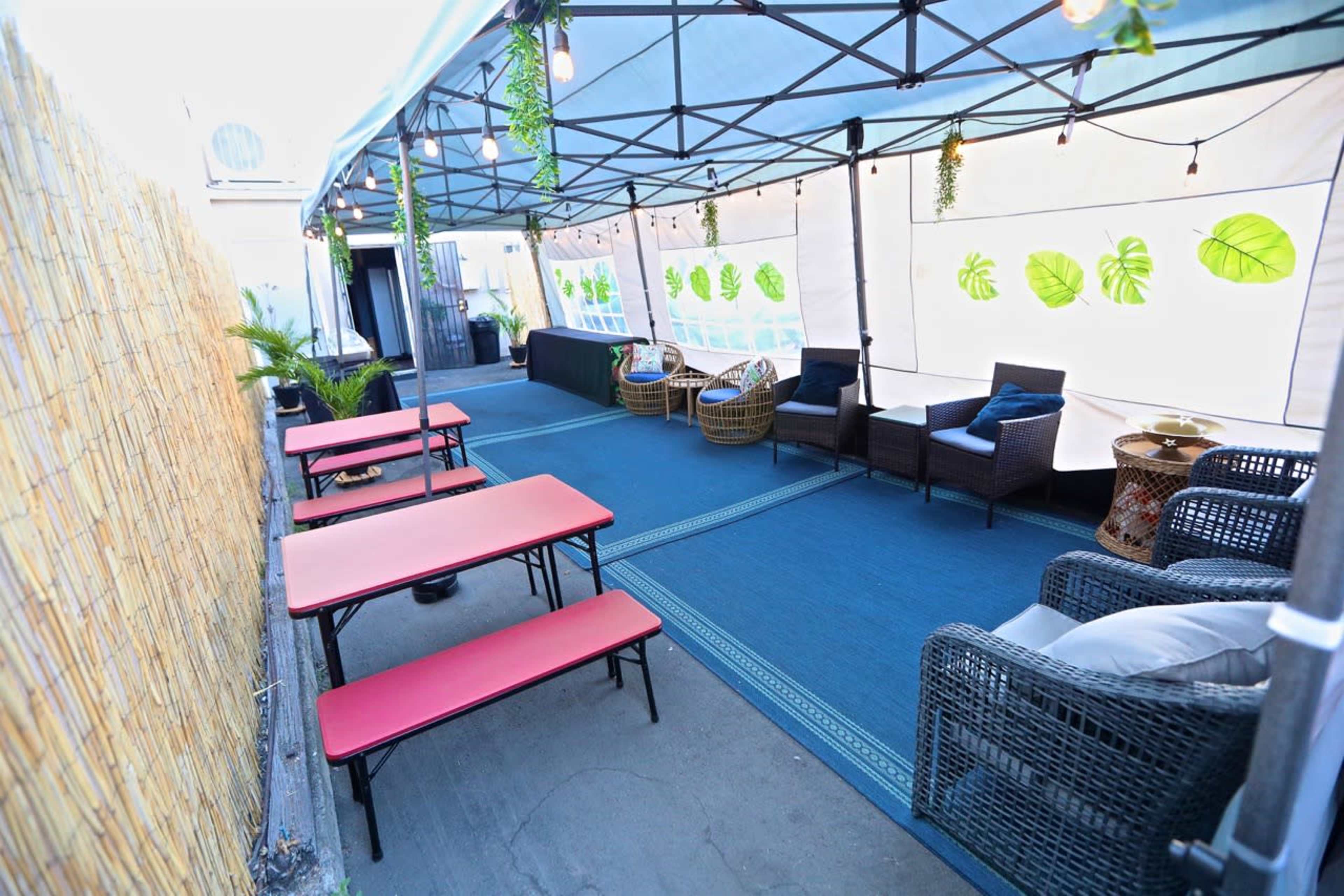 A tented outdoor space features red tables, wicker chairs, and a blue rug, surrounded by bamboo fencing and decorative lighting.