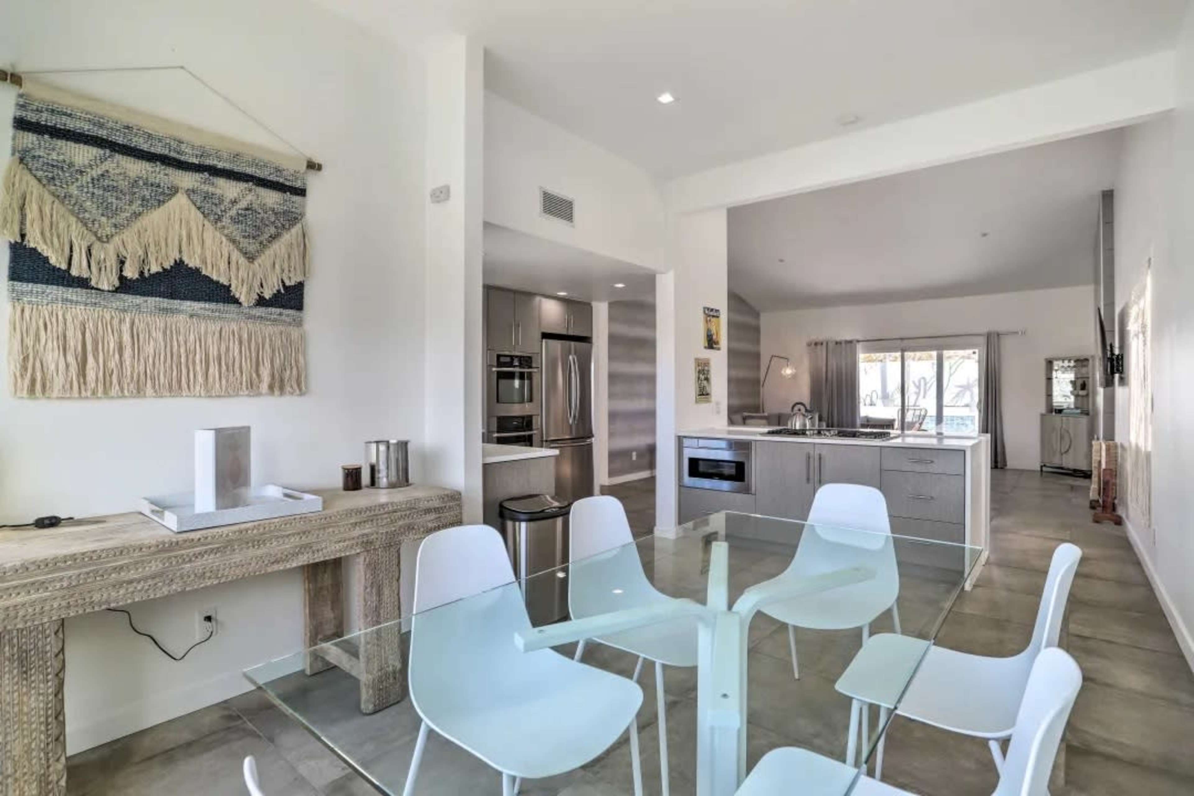 A modern kitchen and dining area features a glass table surrounded by white chairs, with a wooden console table and a stylish wall hanging.