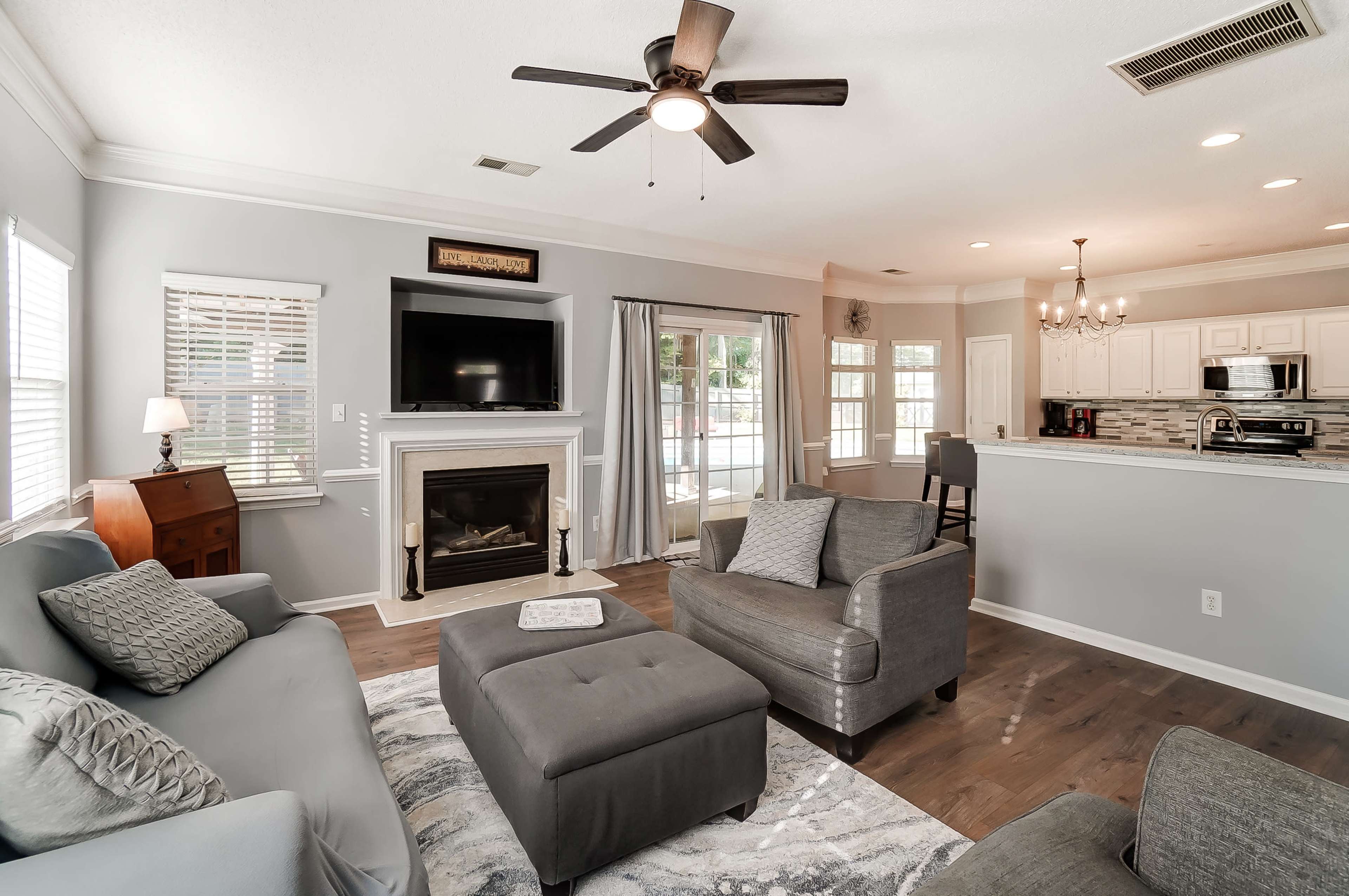 The living room features a gray color scheme with a large sectional sofa, an ottoman, a fireplace, and a television mounted above it, adjacent to an open kitchen area.
