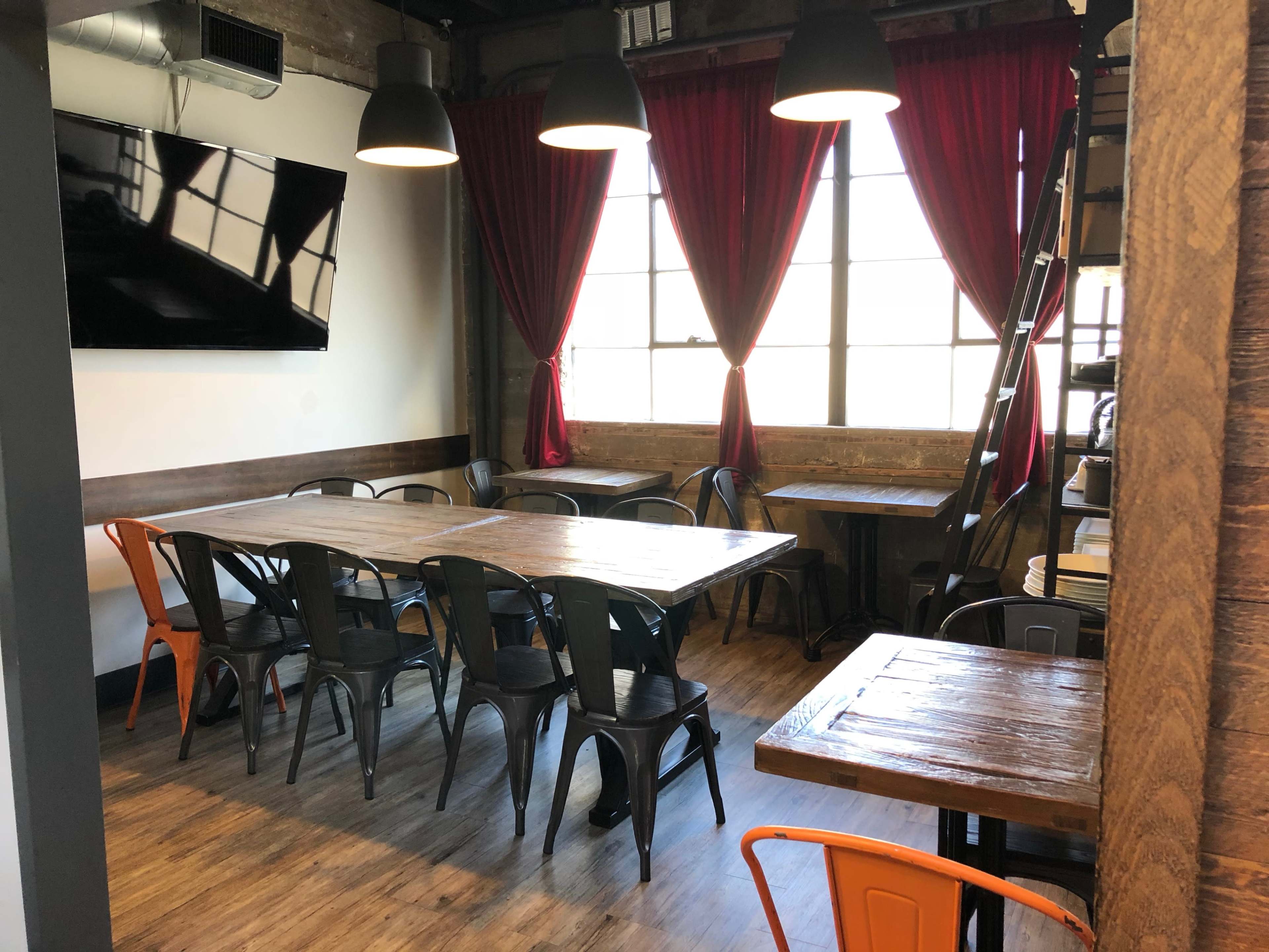 A dining area features wooden tables, metal chairs, and large windows with red curtains, illuminated by hanging lights.