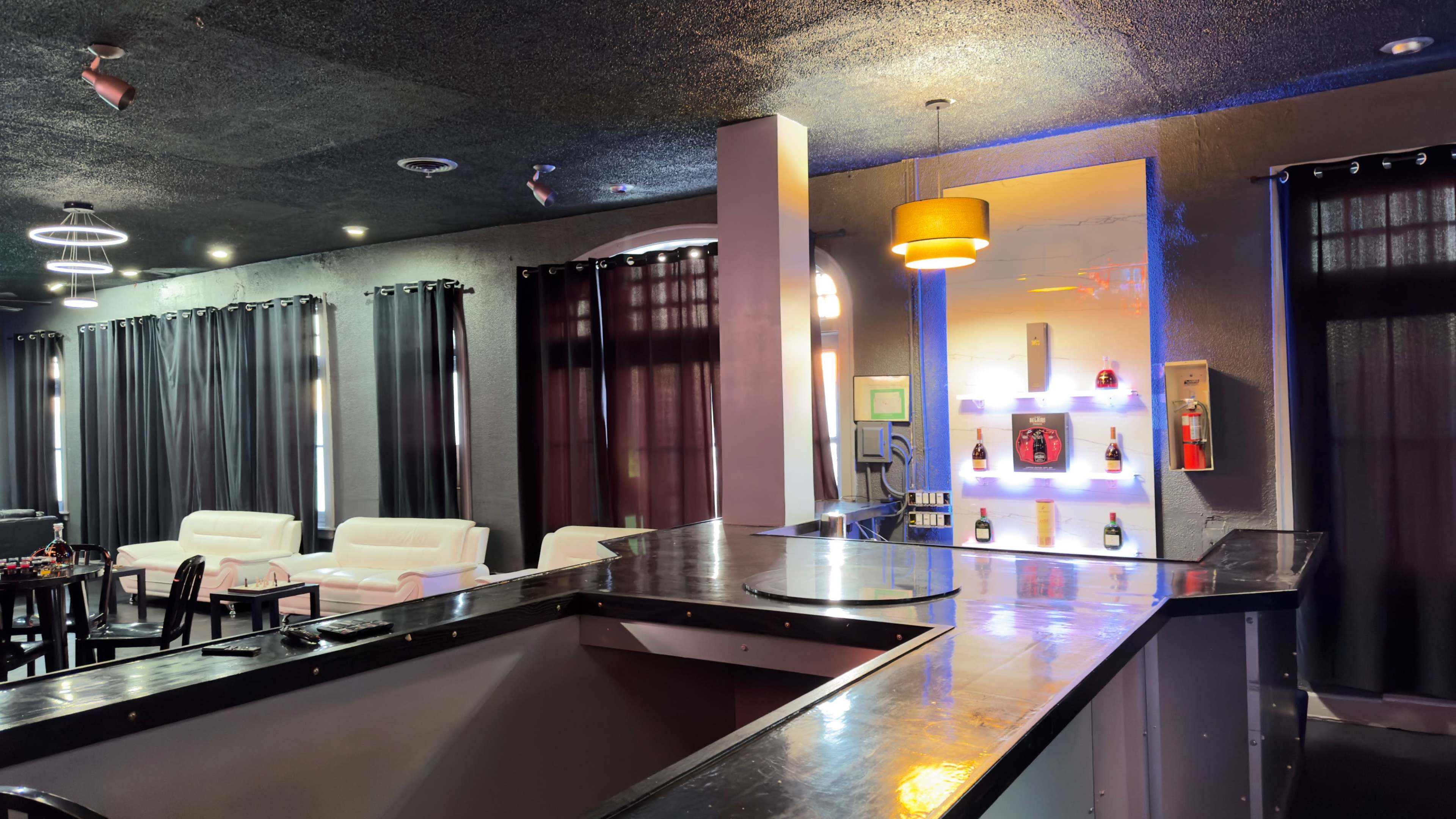 A dimly lit bar area features a sleek black countertop, white leather seating, and a backdrop of dark curtains with a display of bottles illuminated against the wall.