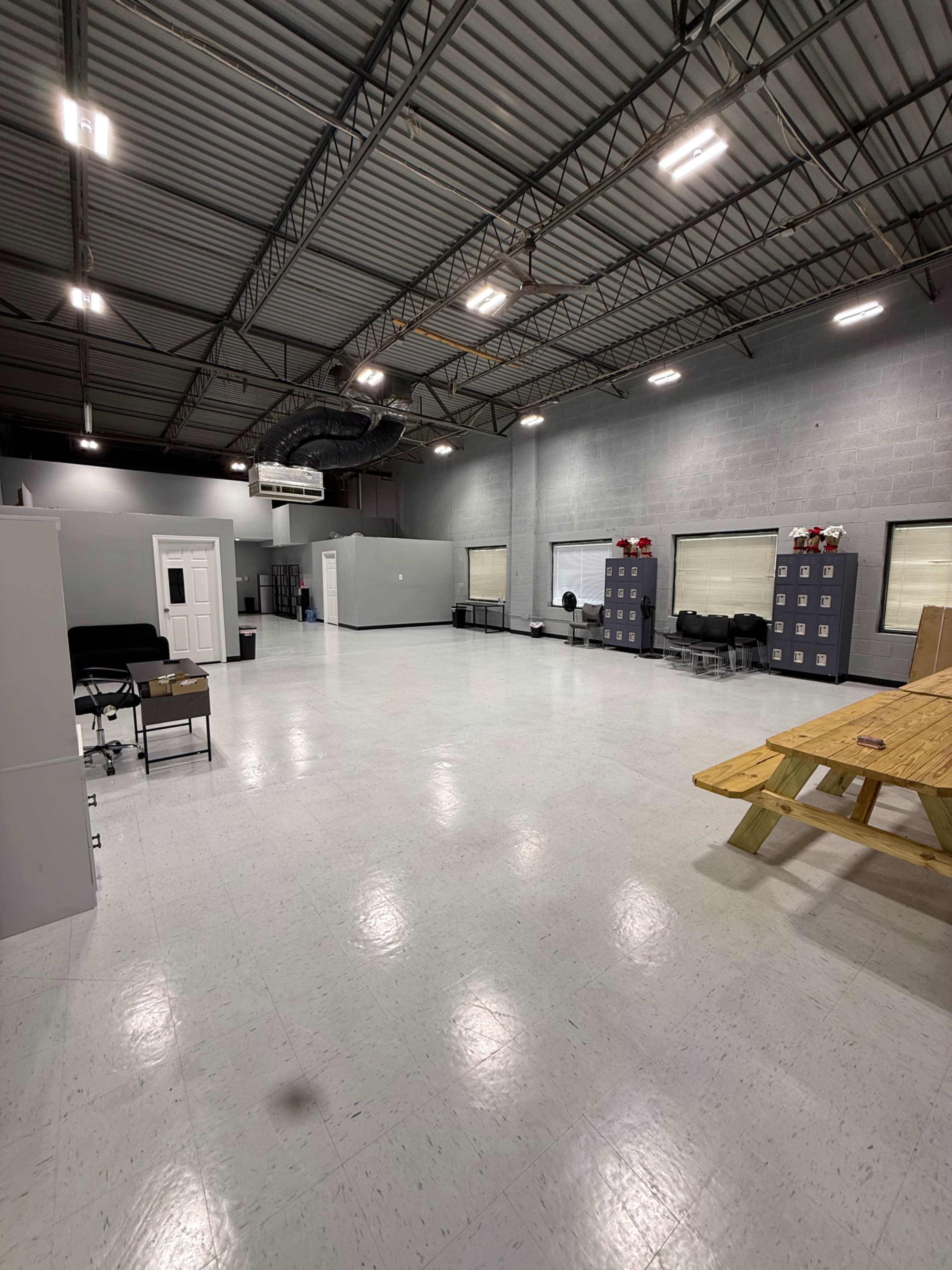 The image shows a spacious, empty room with a high ceiling, gray walls, and a wooden table in the center, along with lockers and a few chairs against the walls.