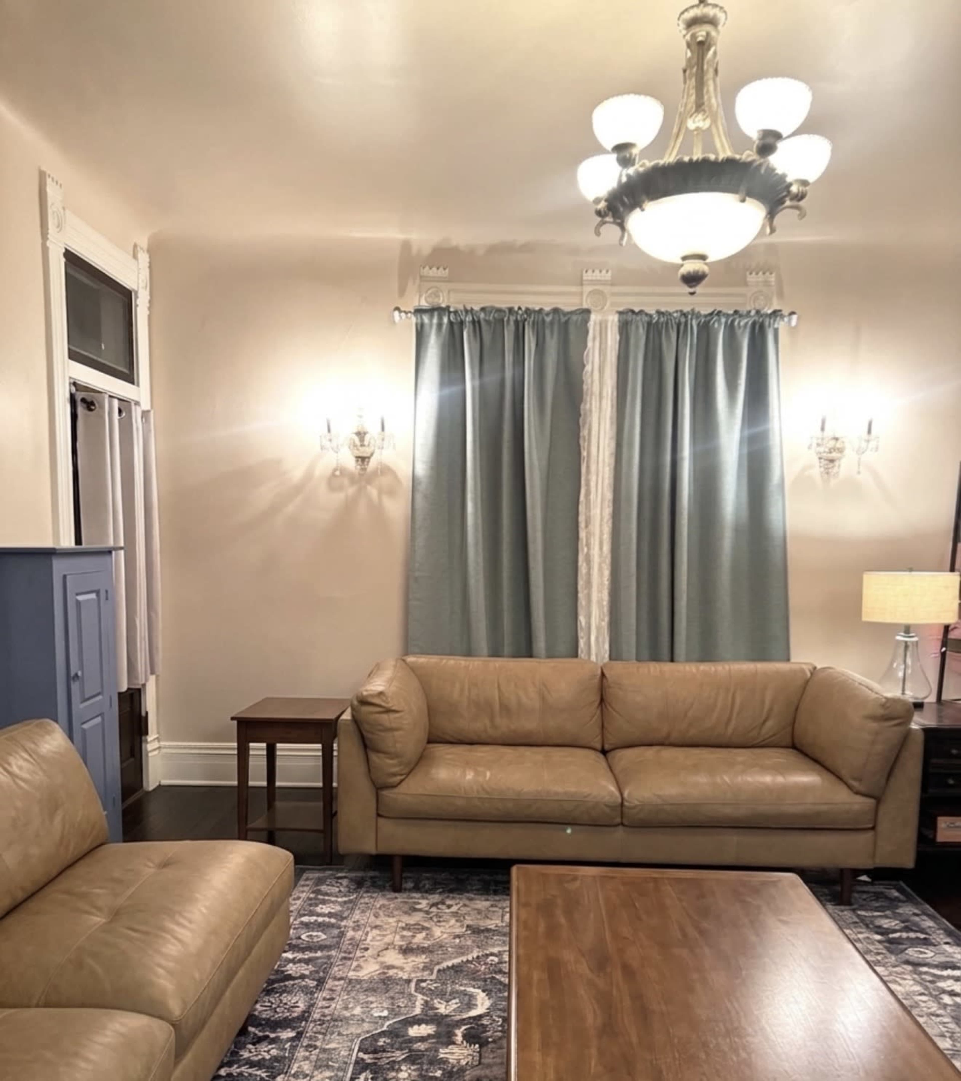 The image shows a living room with two tan leather sofas, a wooden coffee table, and curtains flanking a window, illuminated by a chandelier.