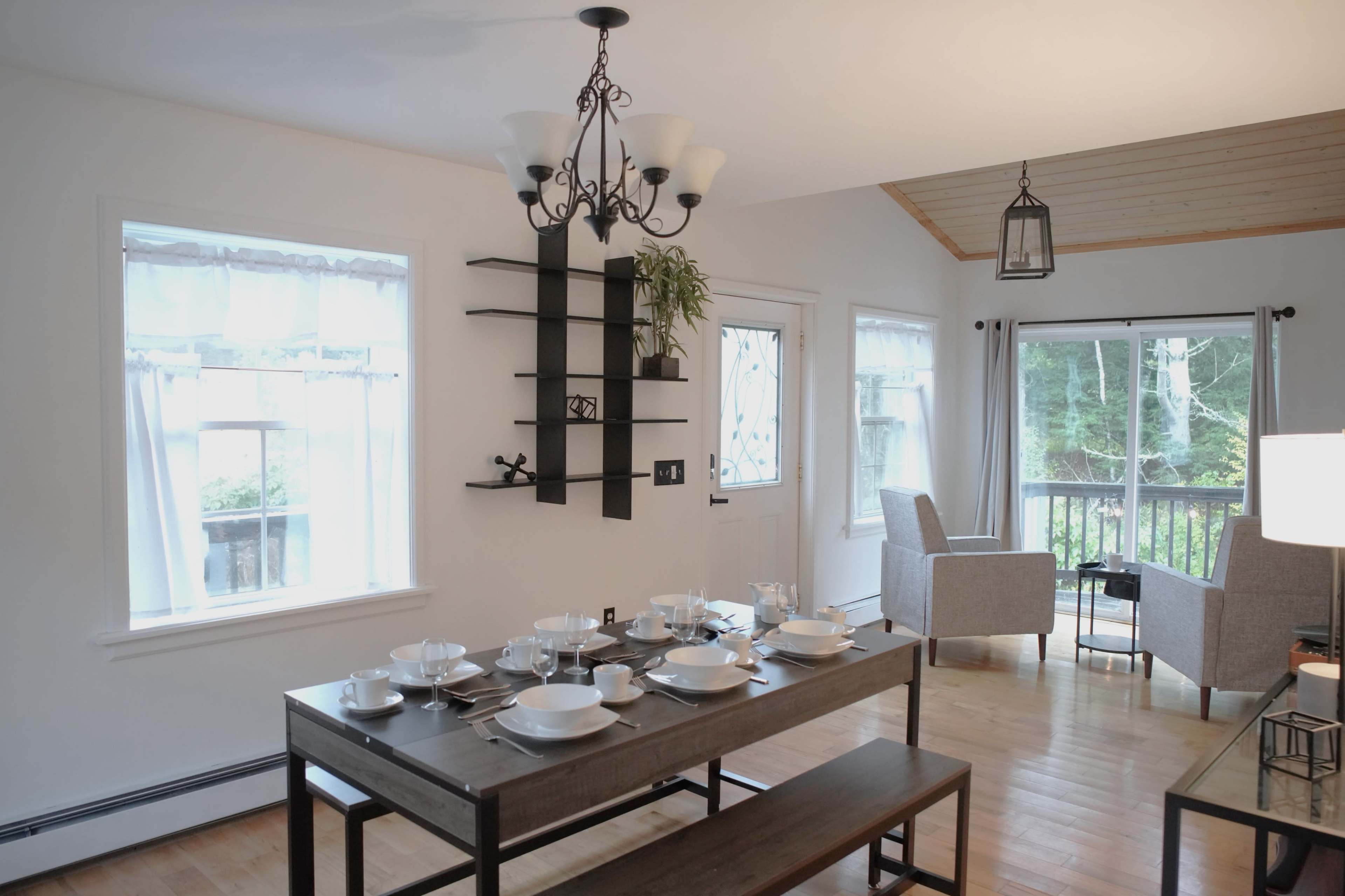 A dining area featuring a wooden table set for a meal, with a chandelier overhead, and a sitting area beside large windows that overlook a deck and greenery outside.