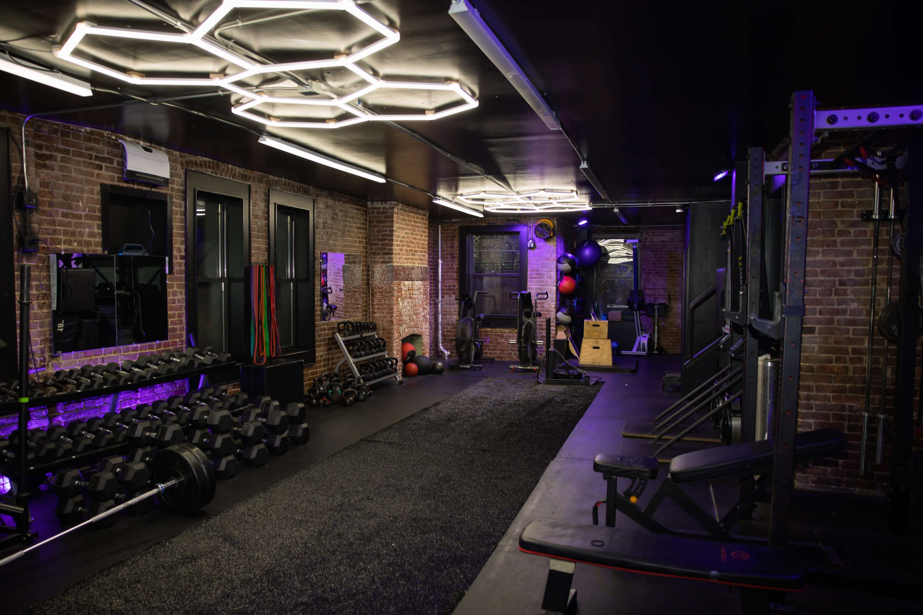 The image shows a modern gym interior with exposed brick walls, various exercise equipment, free weights, and neon lighting.