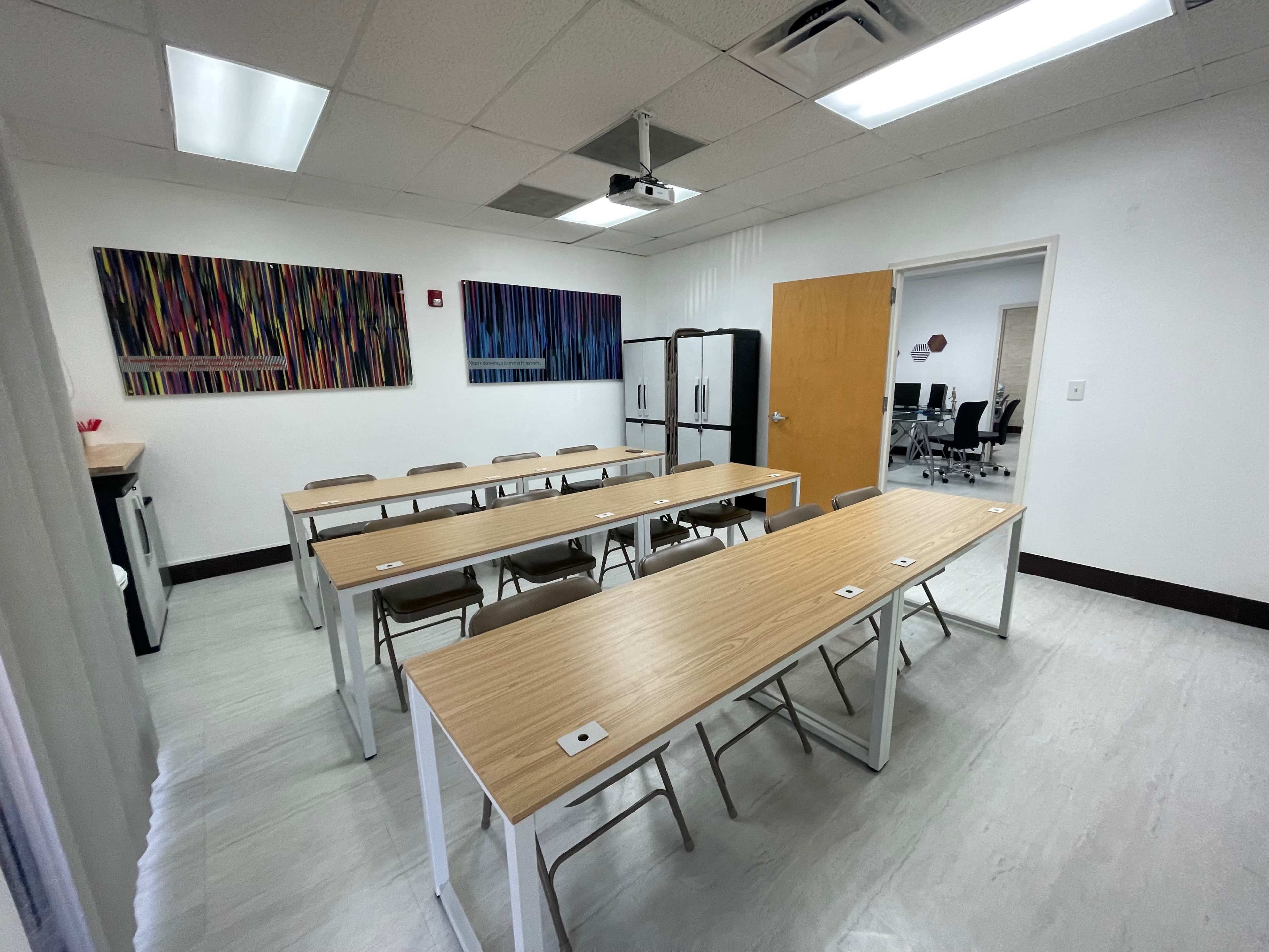 A bright classroom with wooden tables arranged in rows, folding chairs, a refrigerator, and colorful wall art.
