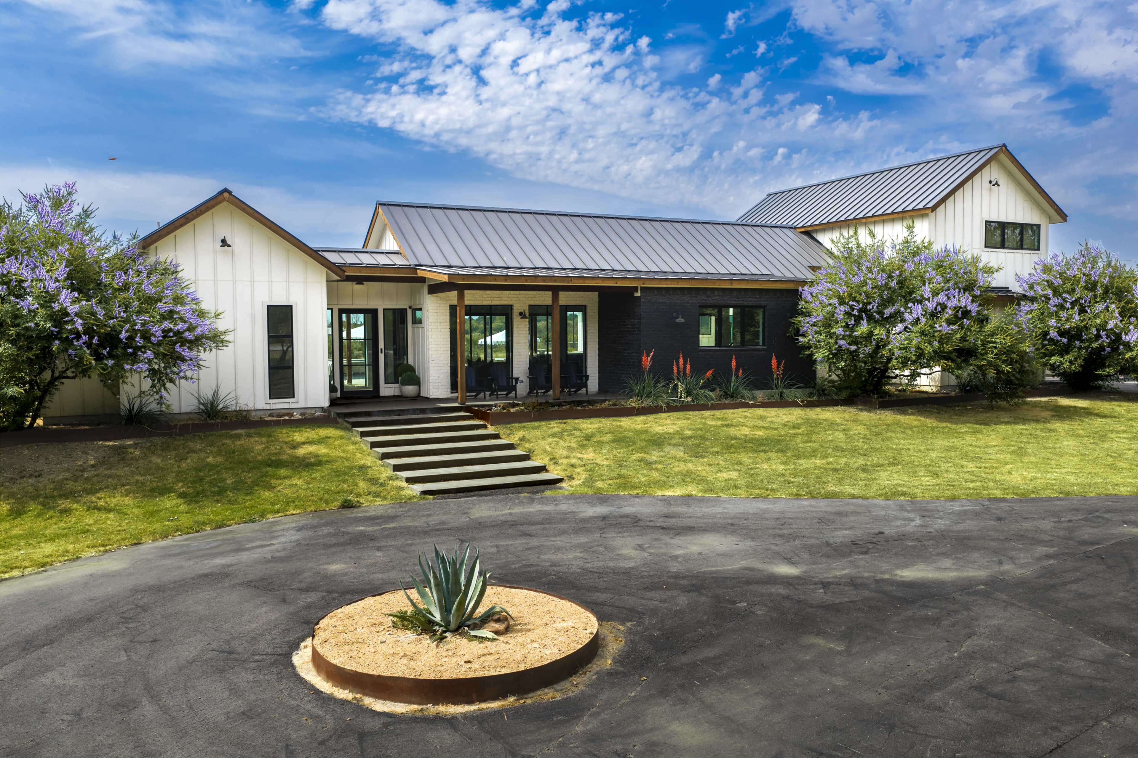 The image shows a modern farmhouse with a metal roof, surrounded by landscaped greenery and flowering shrubs.