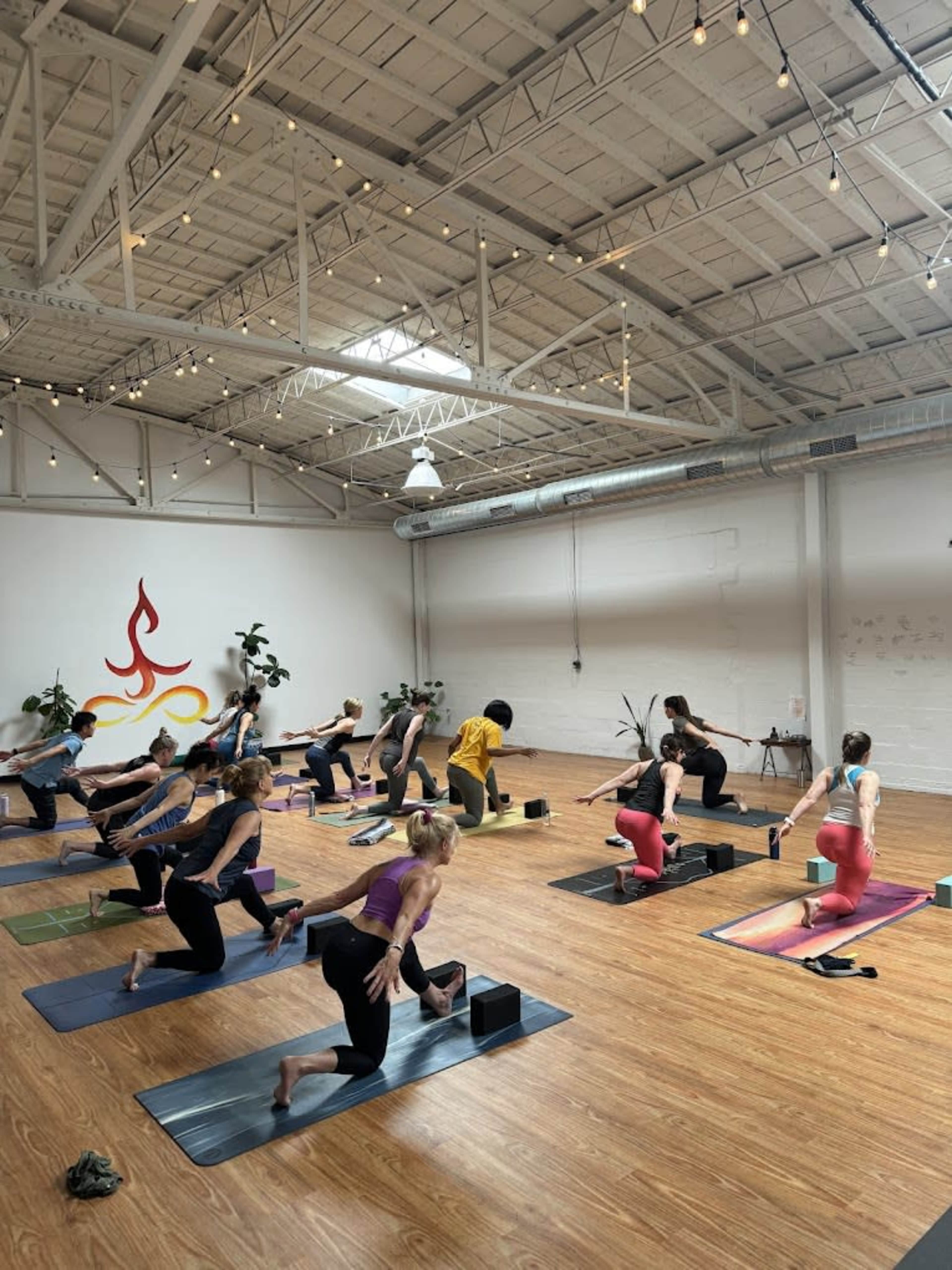 A group of individuals is practicing yoga in a spacious studio with wooden floors and plants, while a colorful logo is visible on the wall.