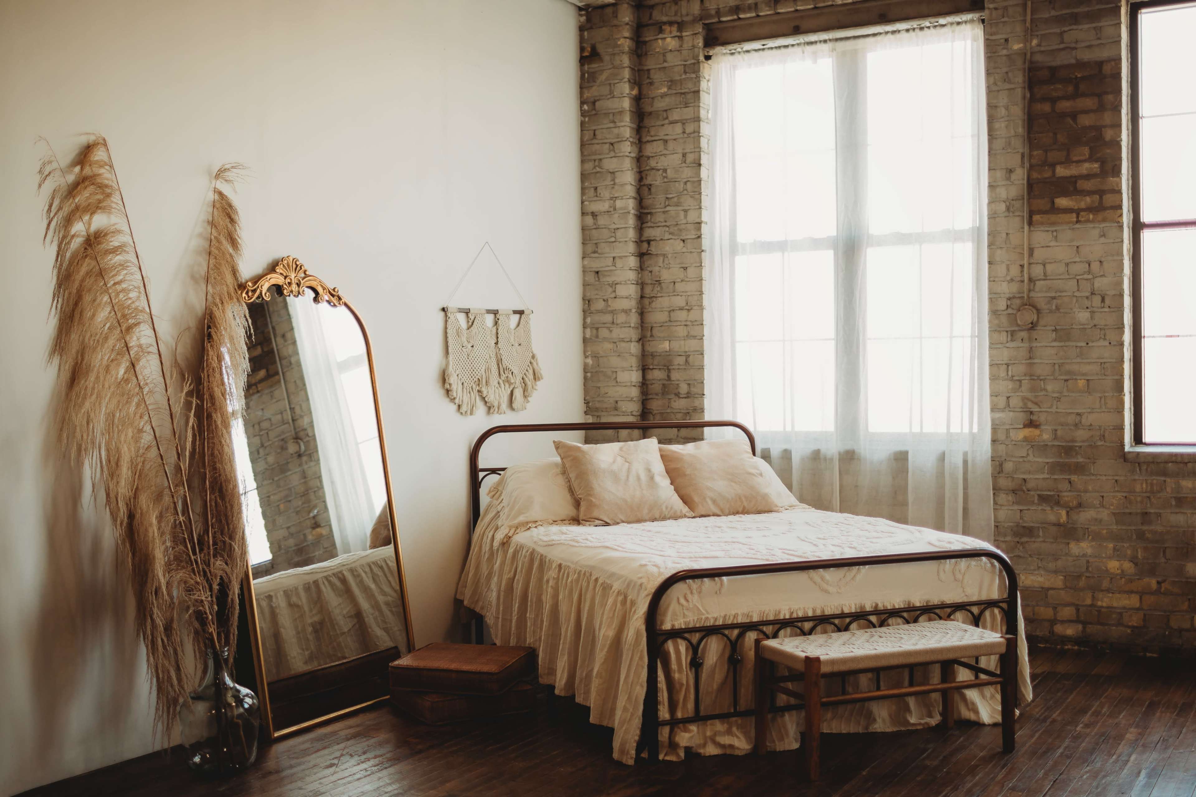 A simple bedroom features a metal frame bed with a light-colored bedspread, a large mirror, and a wooden bench against a wall with exposed brick.