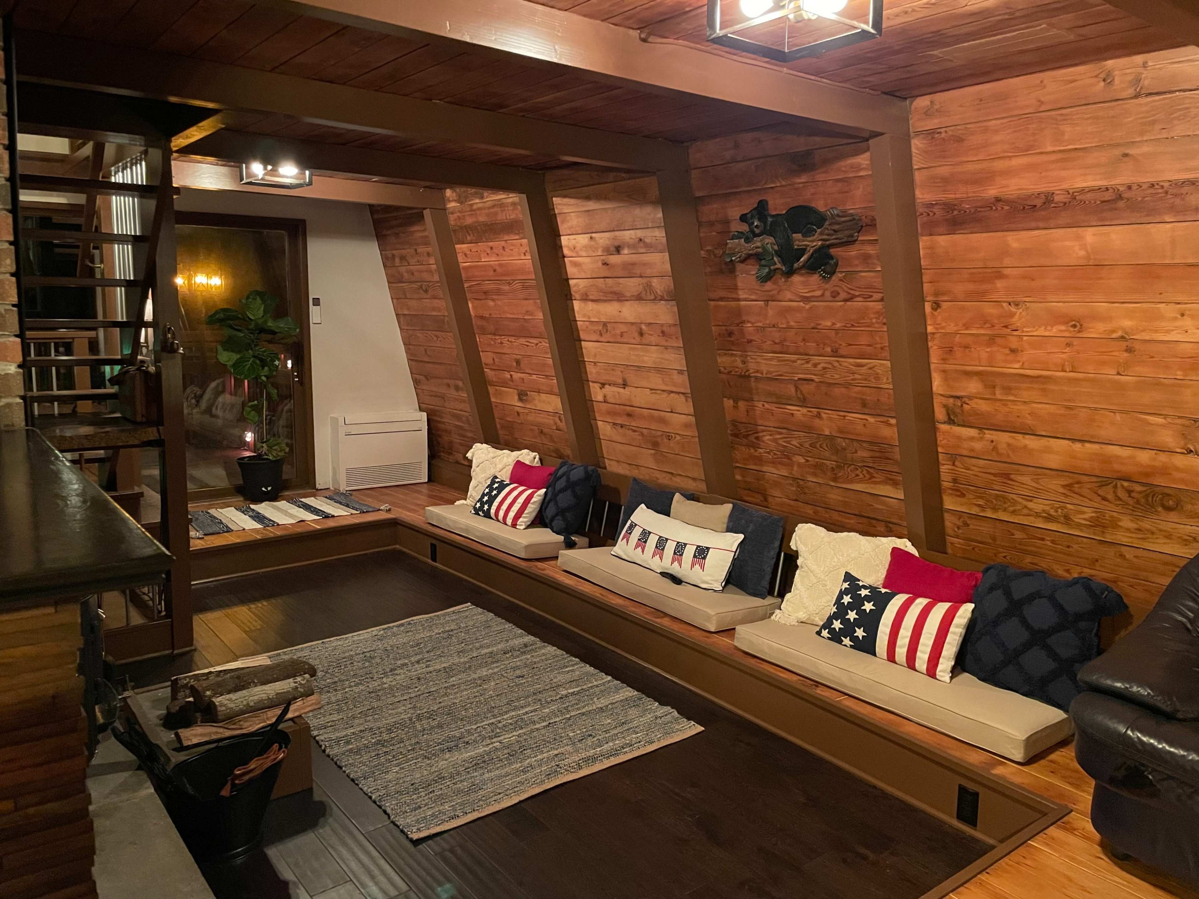 A cozy interior with wooden walls, a long seating area filled with decorative pillows, and a staircase in the background.