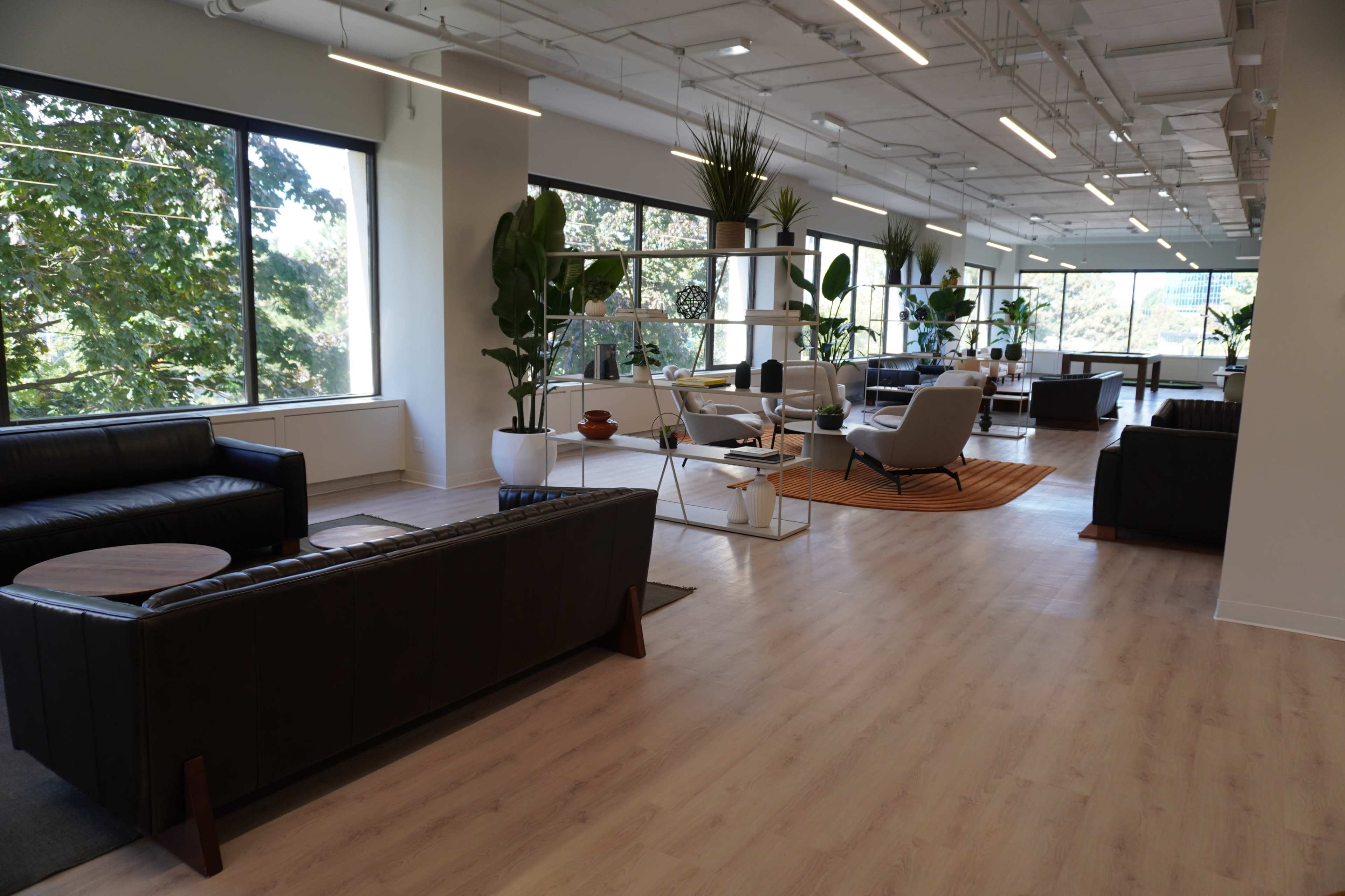The image shows a modern indoor lounge area with large windows, plants, and various seating arrangements, including leather sofas and a coffee table.