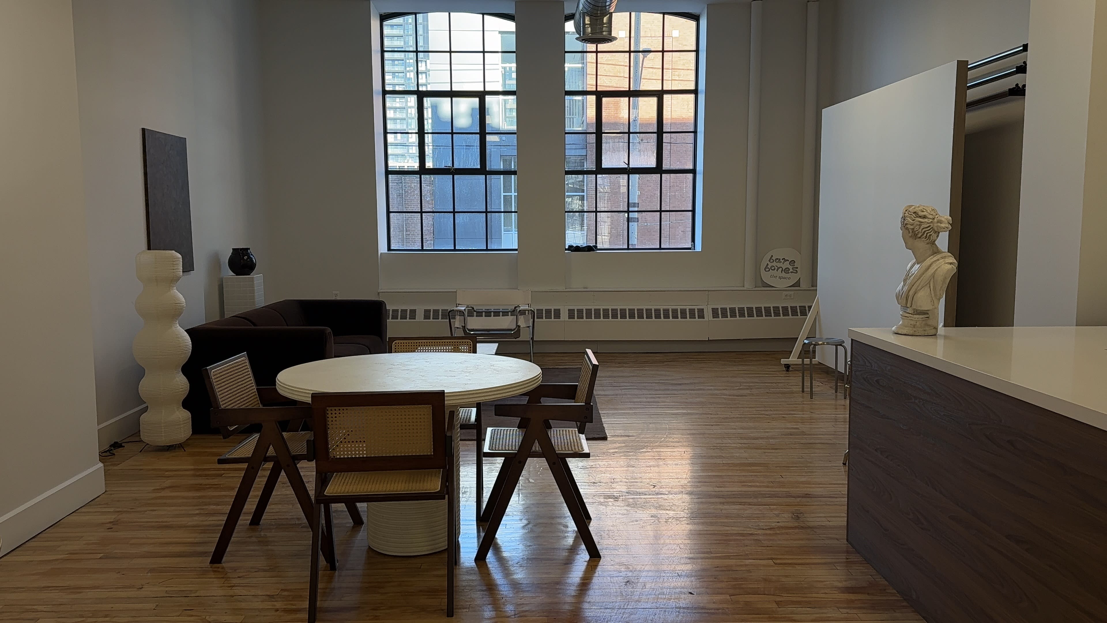 The image shows a spacious interior with a round dining table, wooden chairs, a dark sofa, and large windows allowing natural light to enter.