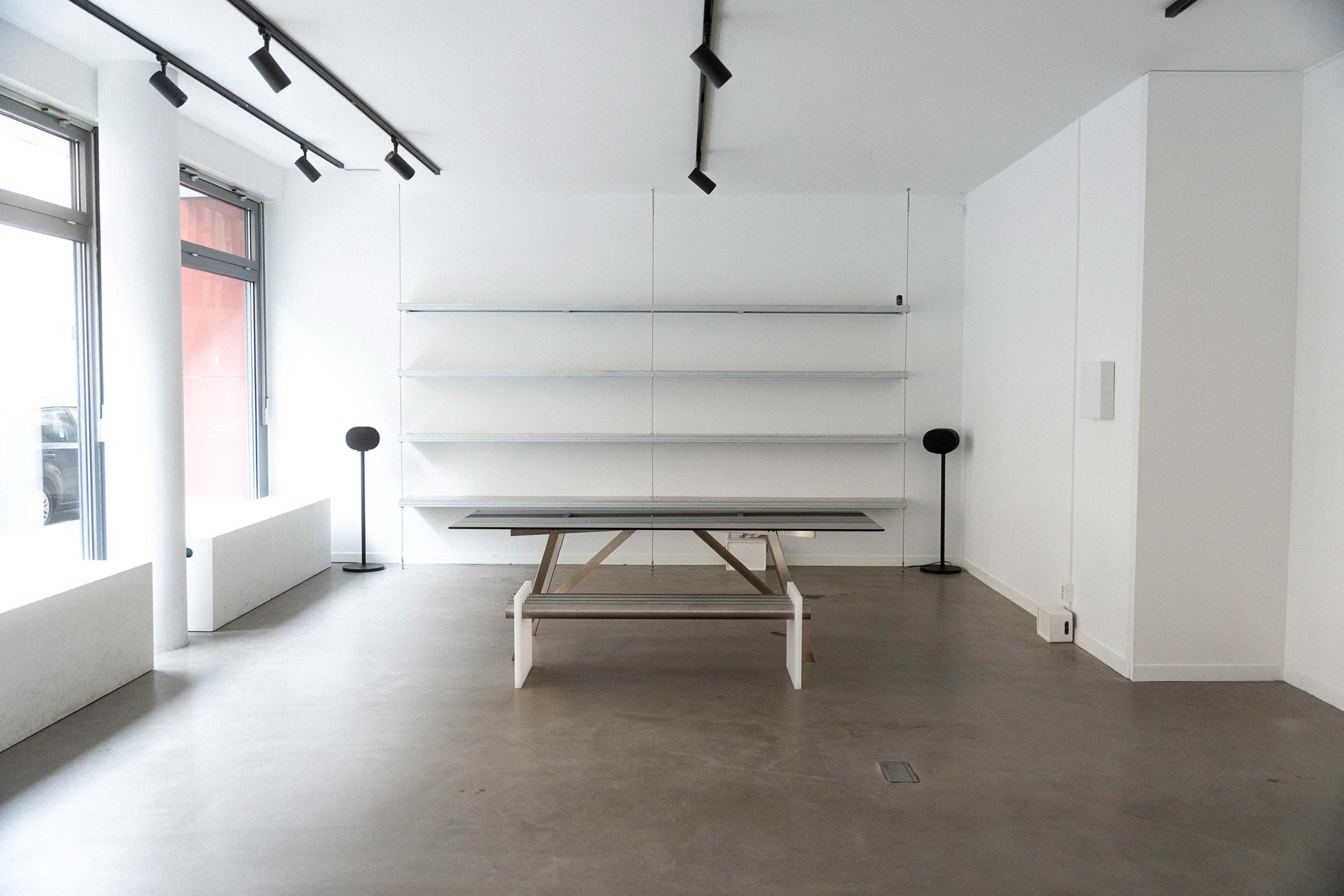 The image shows a minimalist interior space with a central table, two floor lamps, and empty shelves against white walls.