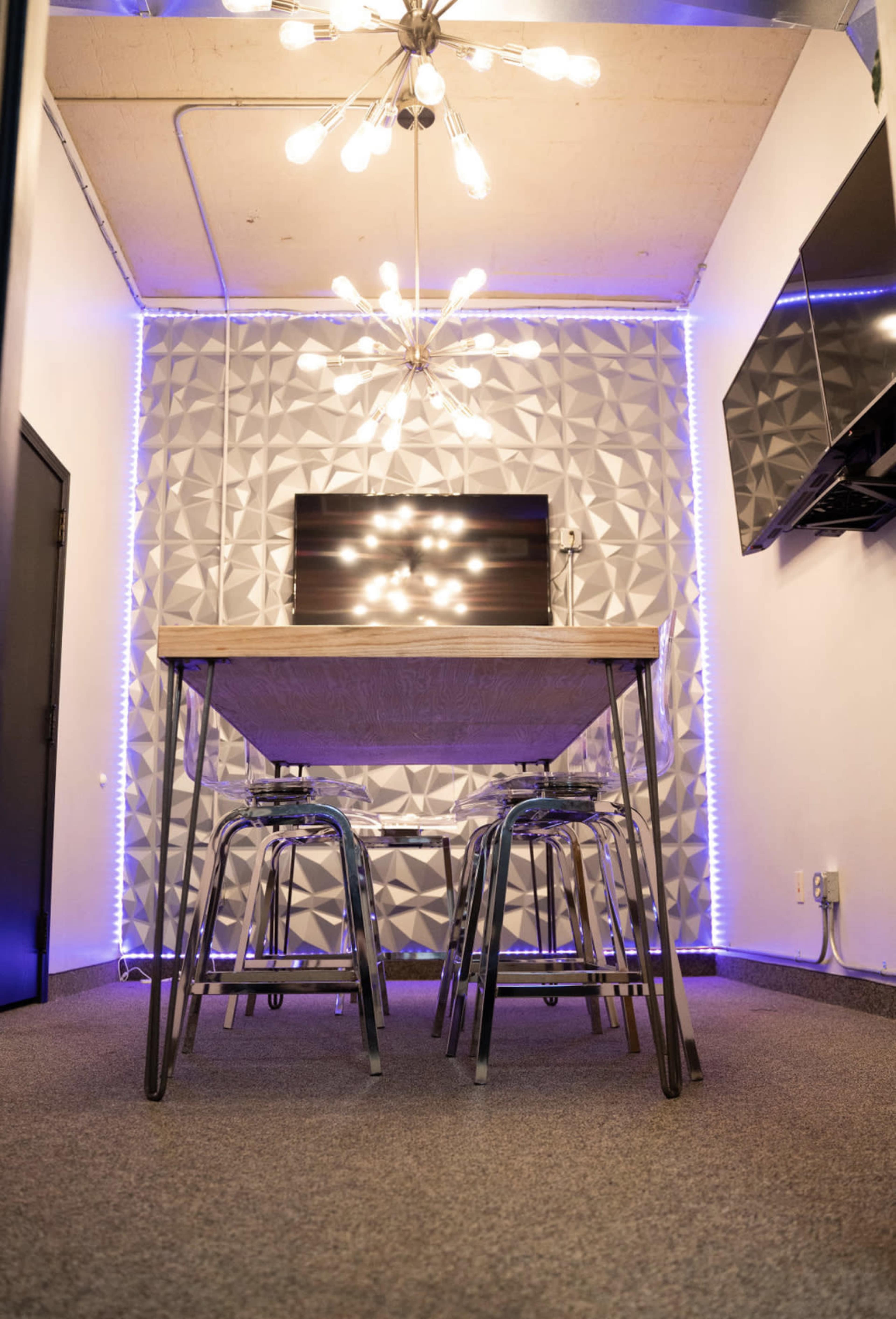 The image shows a modern workspace featuring a wooden table with metal stools, illuminated by a chandelier and surrounded by textured wall panels with purple lighting.