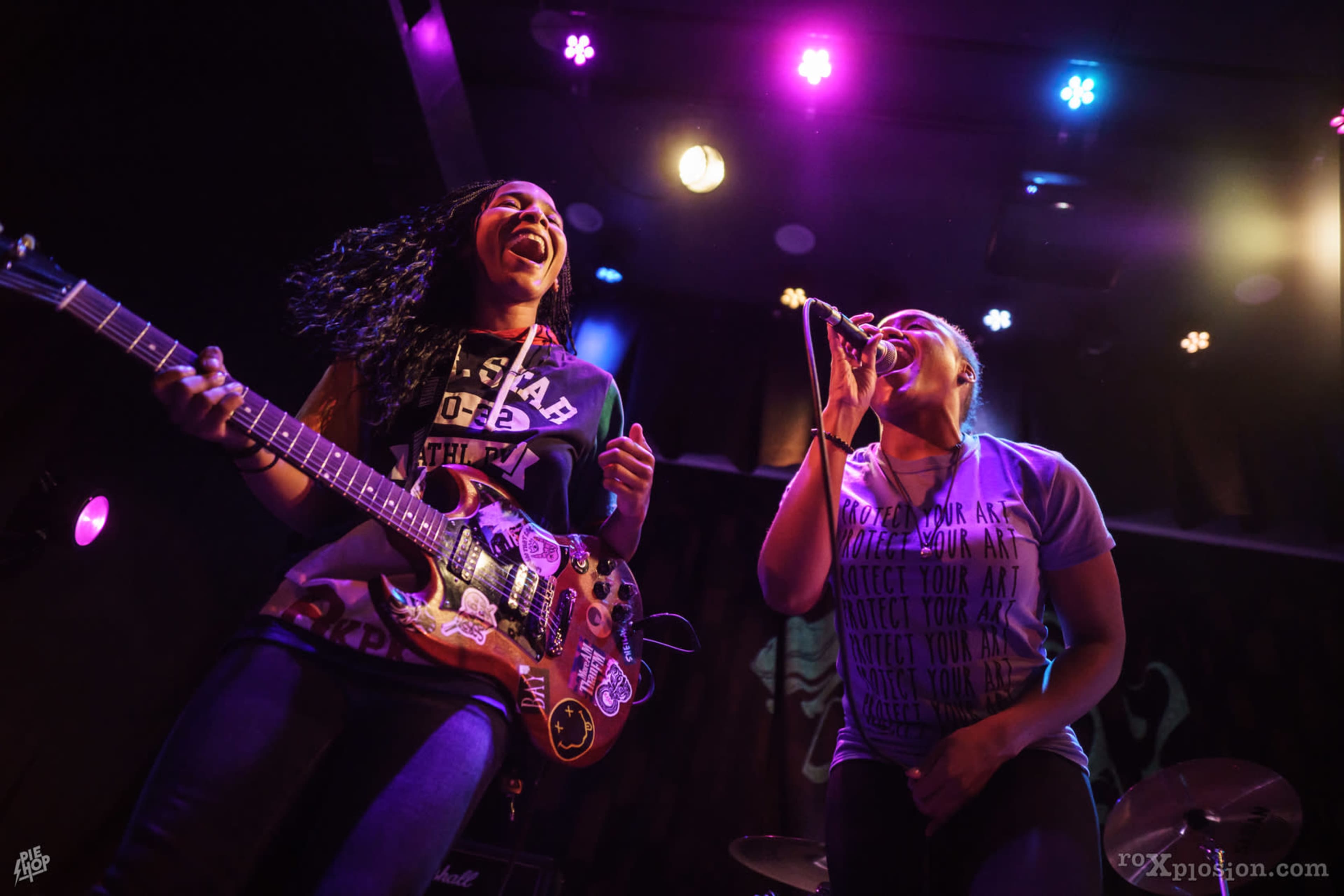 Two women perform on stage, one playing an electric guitar and the other singing into a microphone, with colorful stage lights illuminating the scene.