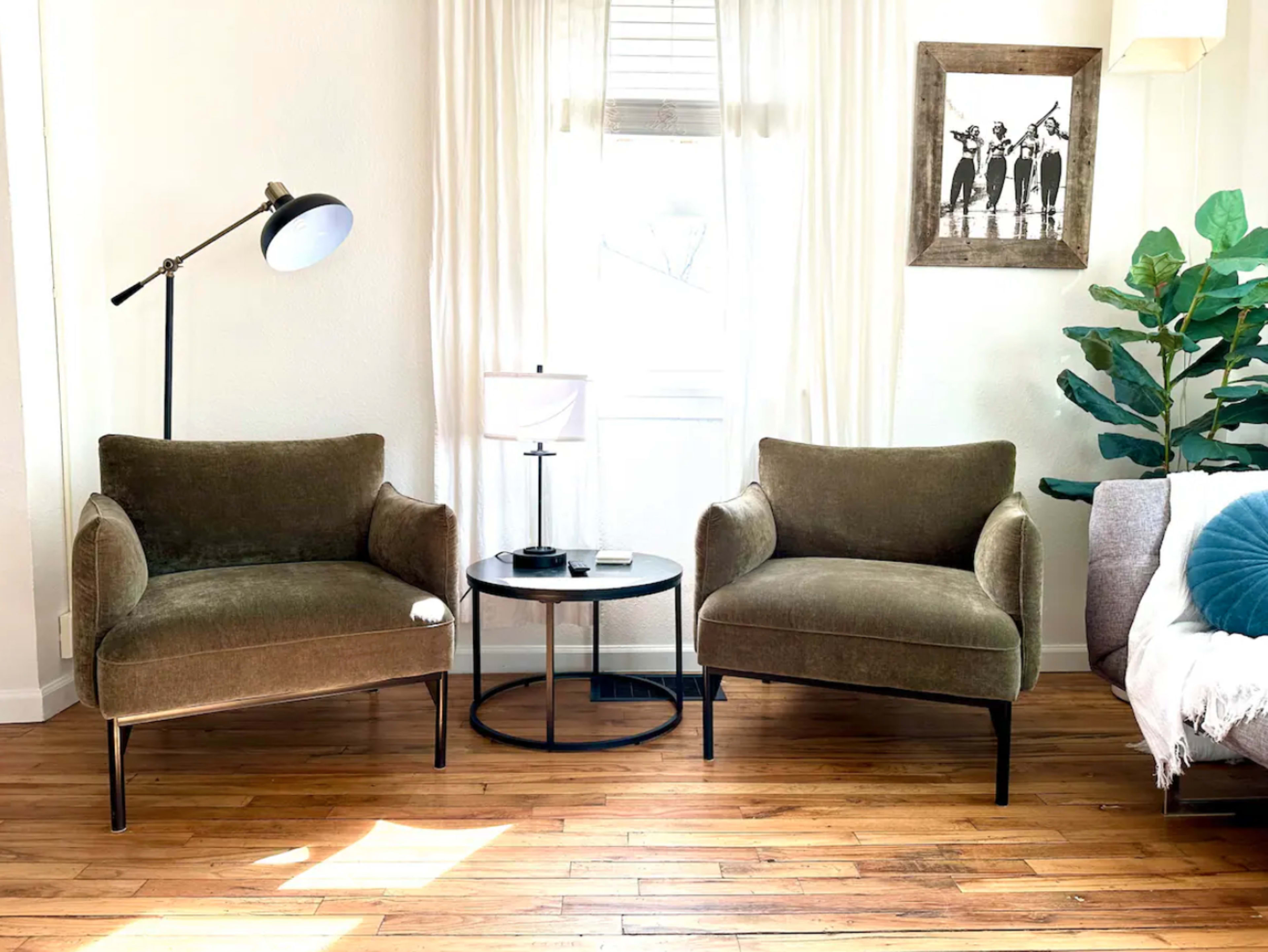 Two green armchairs next to a round black side table with a lamp and a plant, set in a brightly lit room with white curtains and a framed picture on the wall.