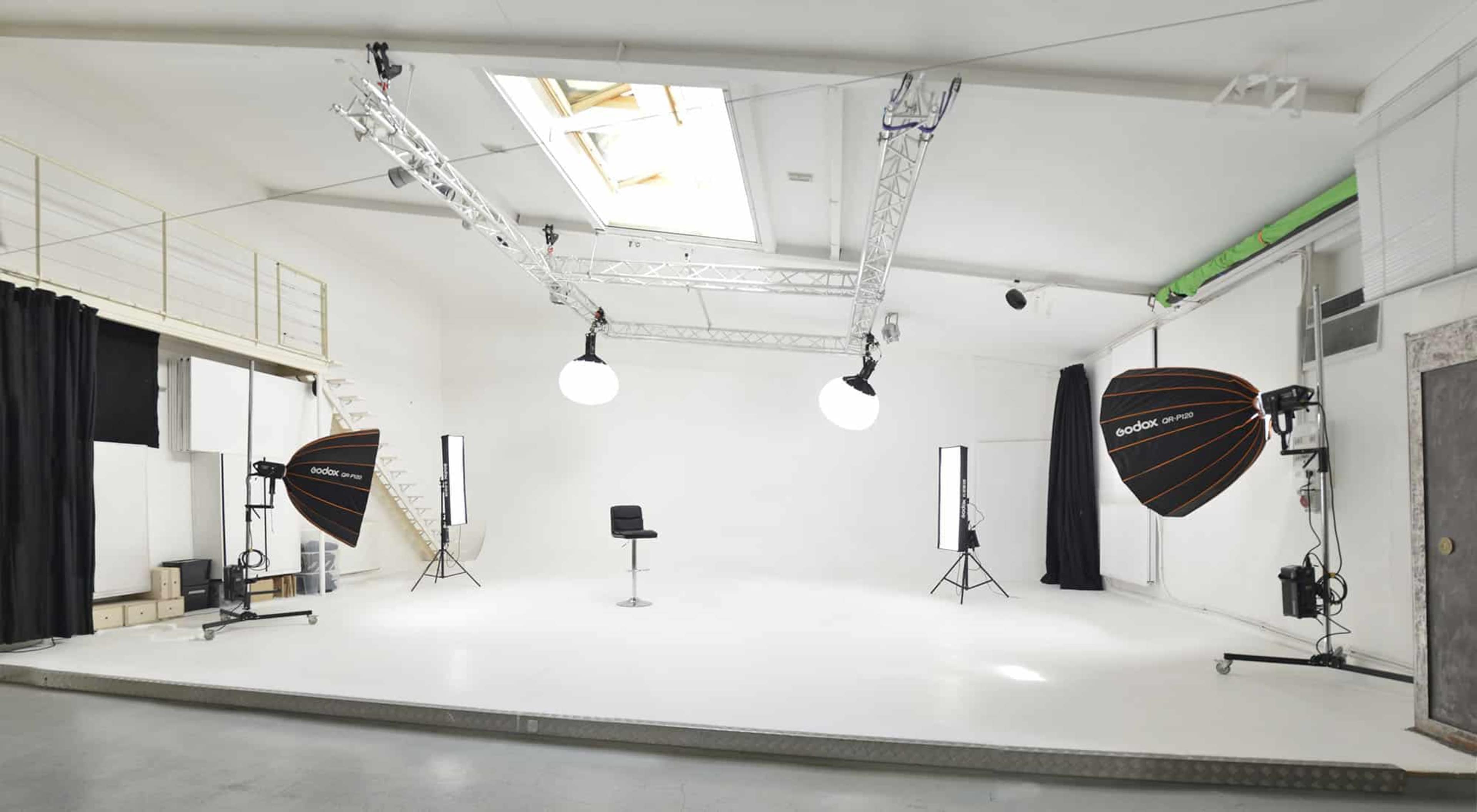 A photography studio features a large white backdrop with multiple lighting setups, including softboxes and a high chair in the center.