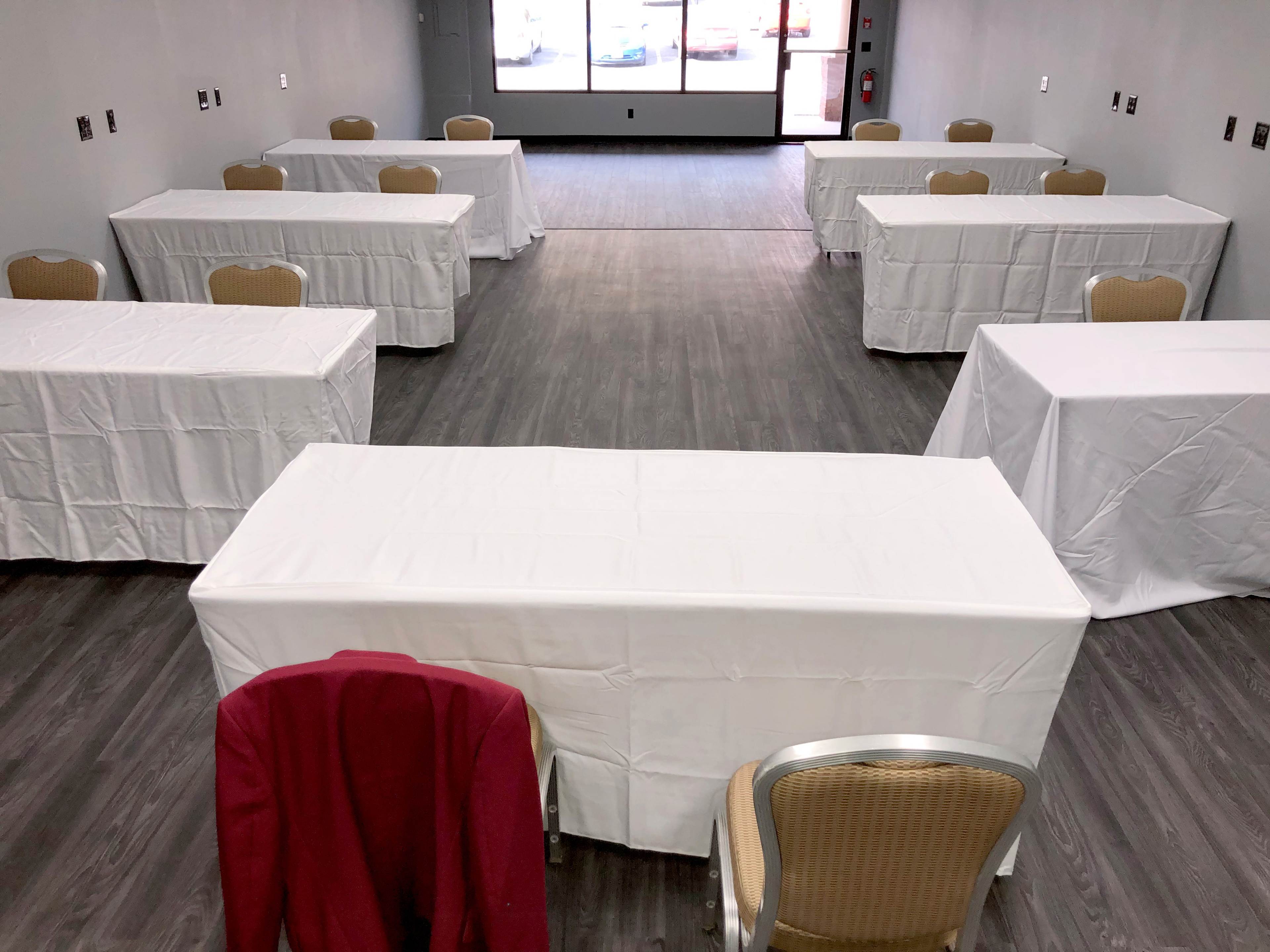A spacious room with several tables covered in white tablecloths arranged in a grid pattern, with a red jacket draped over one chair.