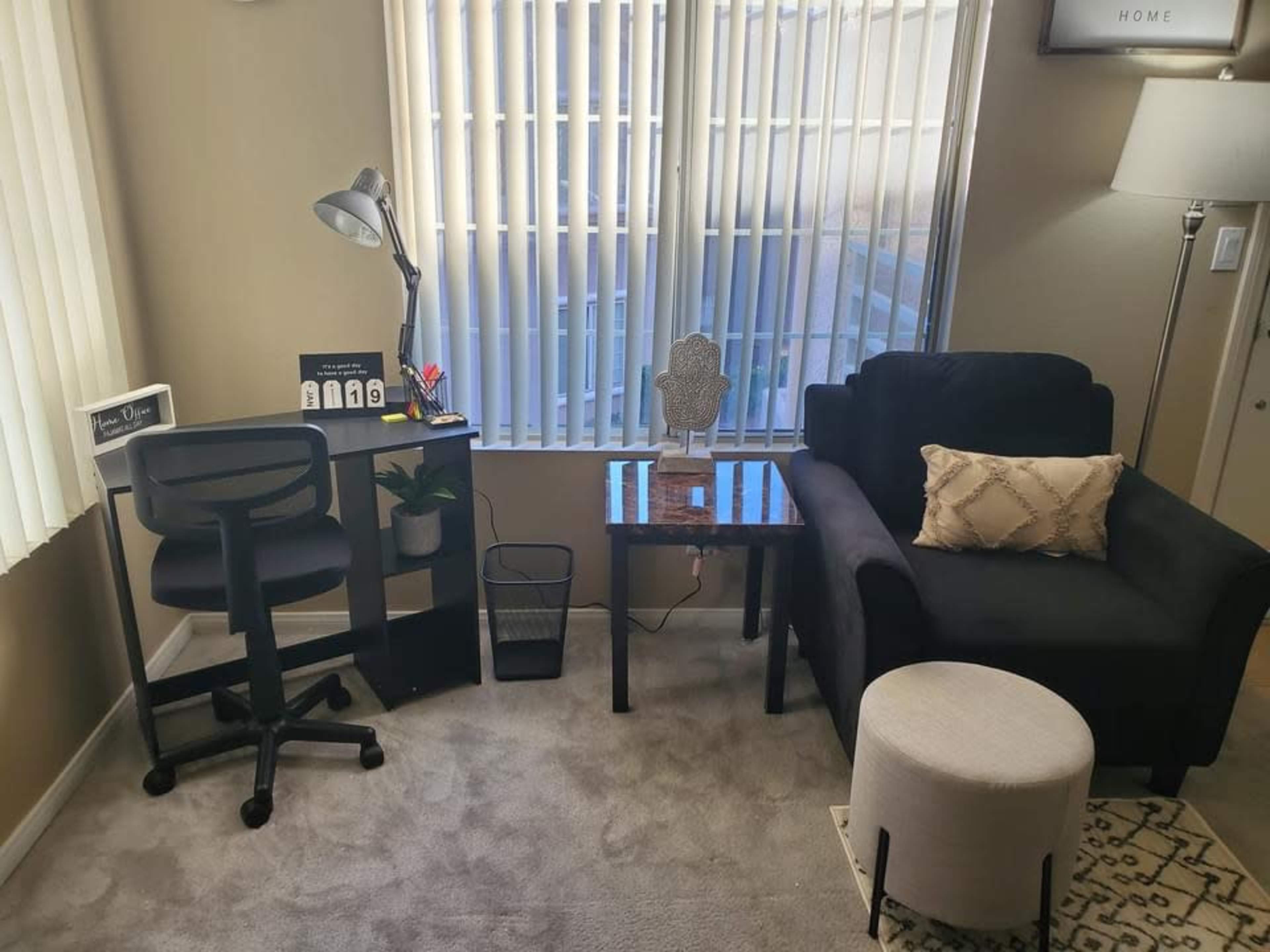 The living space features a black chair, a small table with a decorative item, a white ottoman, a desk with a lamp, and large windows letting in natural light.