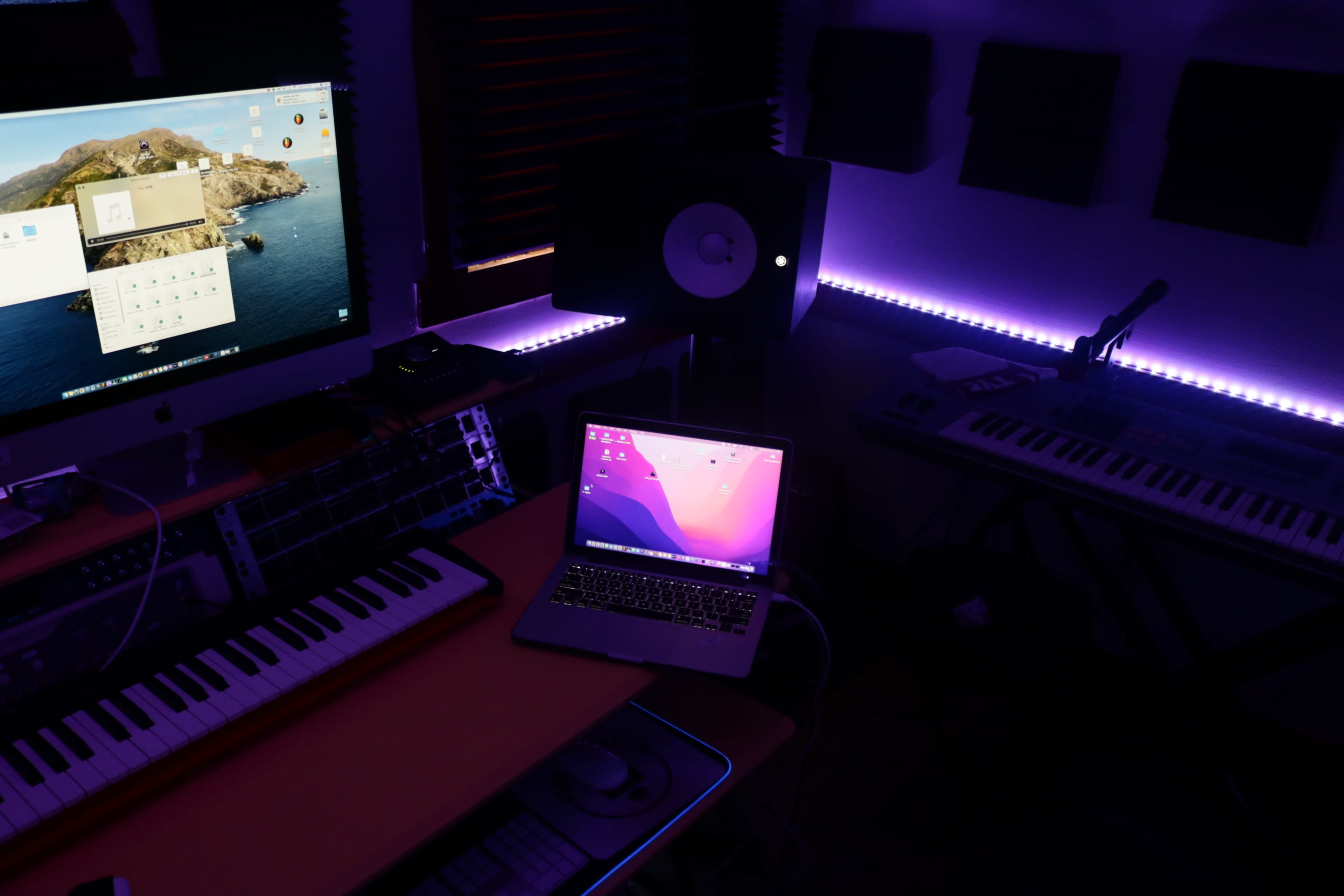 A music production studio with a desktop monitor, a laptop, keyboards, and ambient lighting.