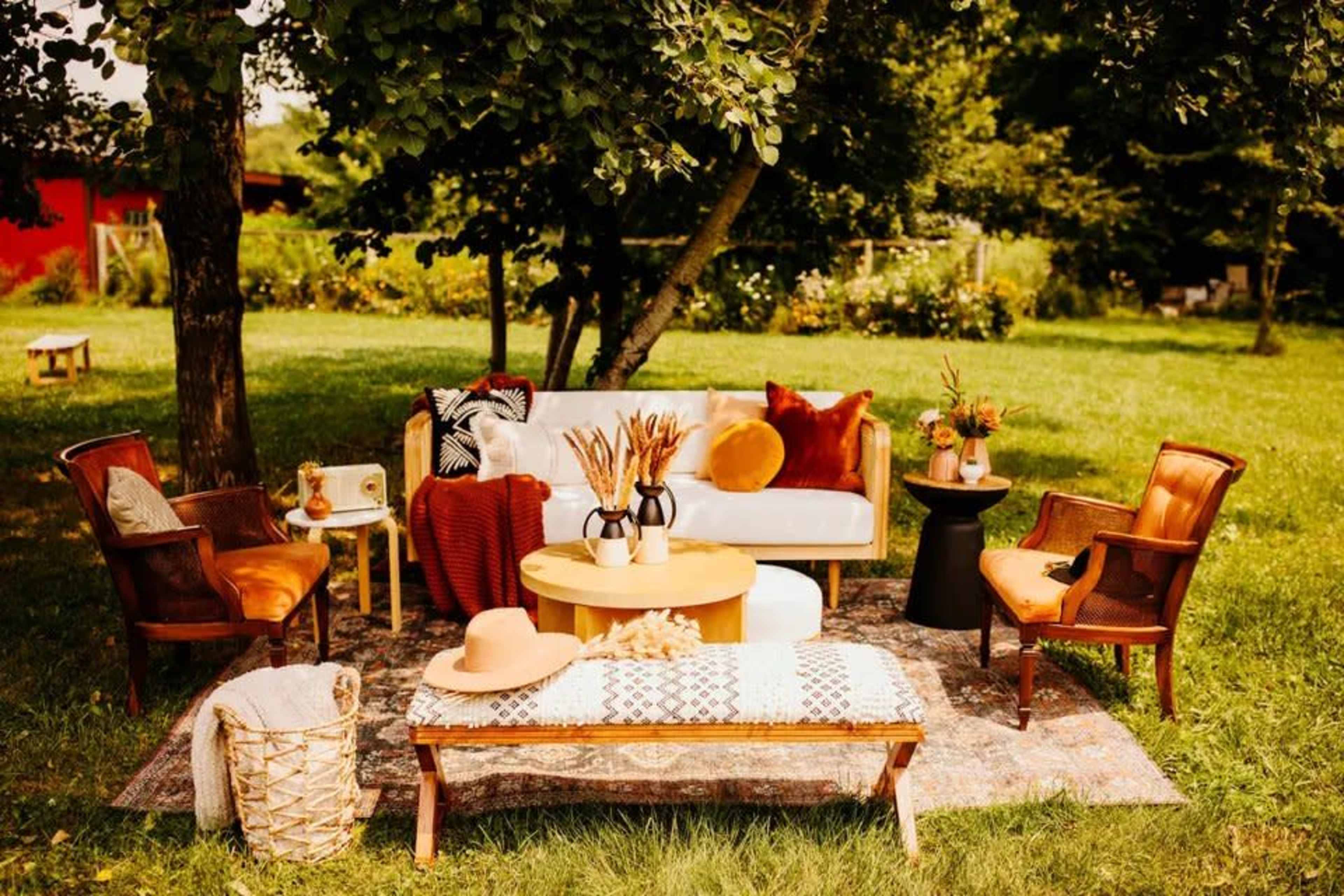 A cozy outdoor seating area features a white sofa, wooden chairs, and a round coffee table surrounded by green grass and trees.