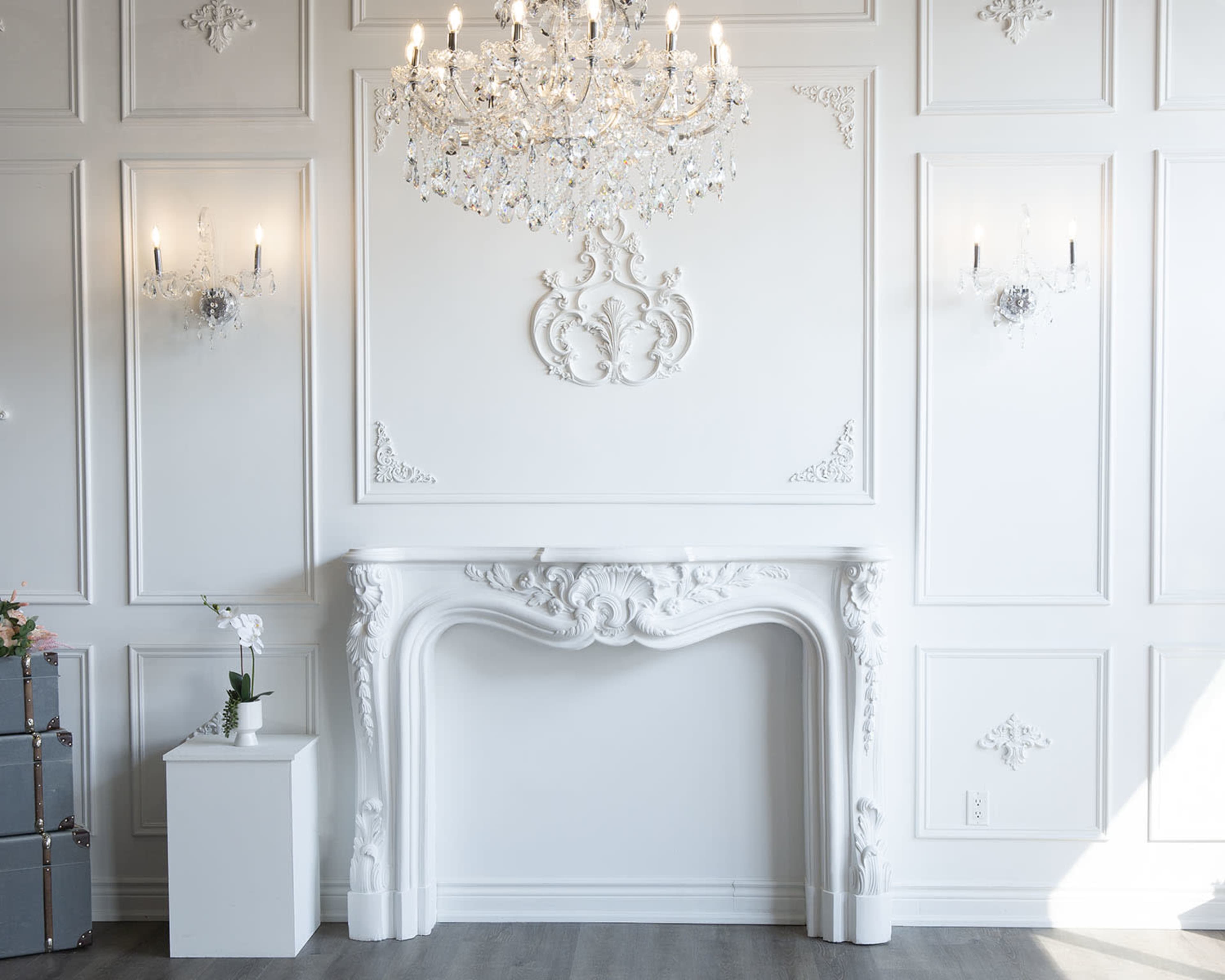 A white wall with intricate moldings, a decorative fireplace, and a crystal chandelier above.